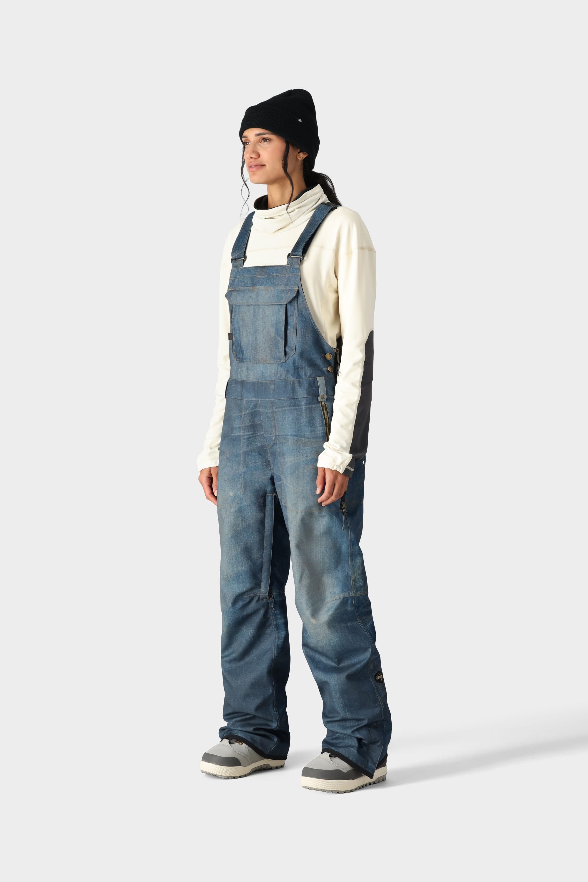 Someone wearing the 686 Womens Black Magic 20K Bib by 686 stands turned slightly to the side against a plain light gray background, paired with a black beanie and white long-sleeve shirt.