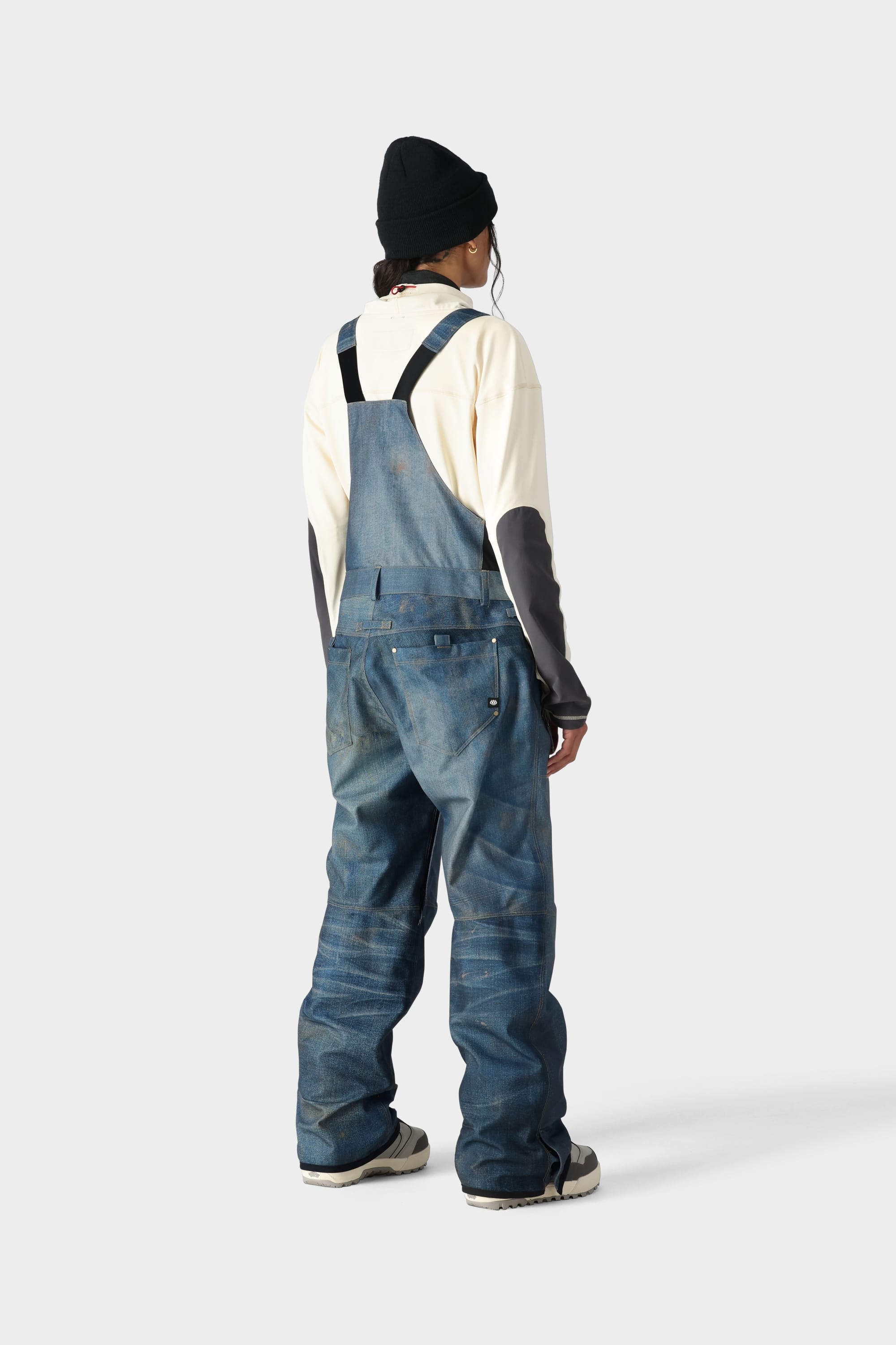A person stands facing away in the 686 Womens Black Magic 20K Bib by 686, styled with a black beanie, cream long-sleeve shirt, and white sneakers against a plain light gray background.