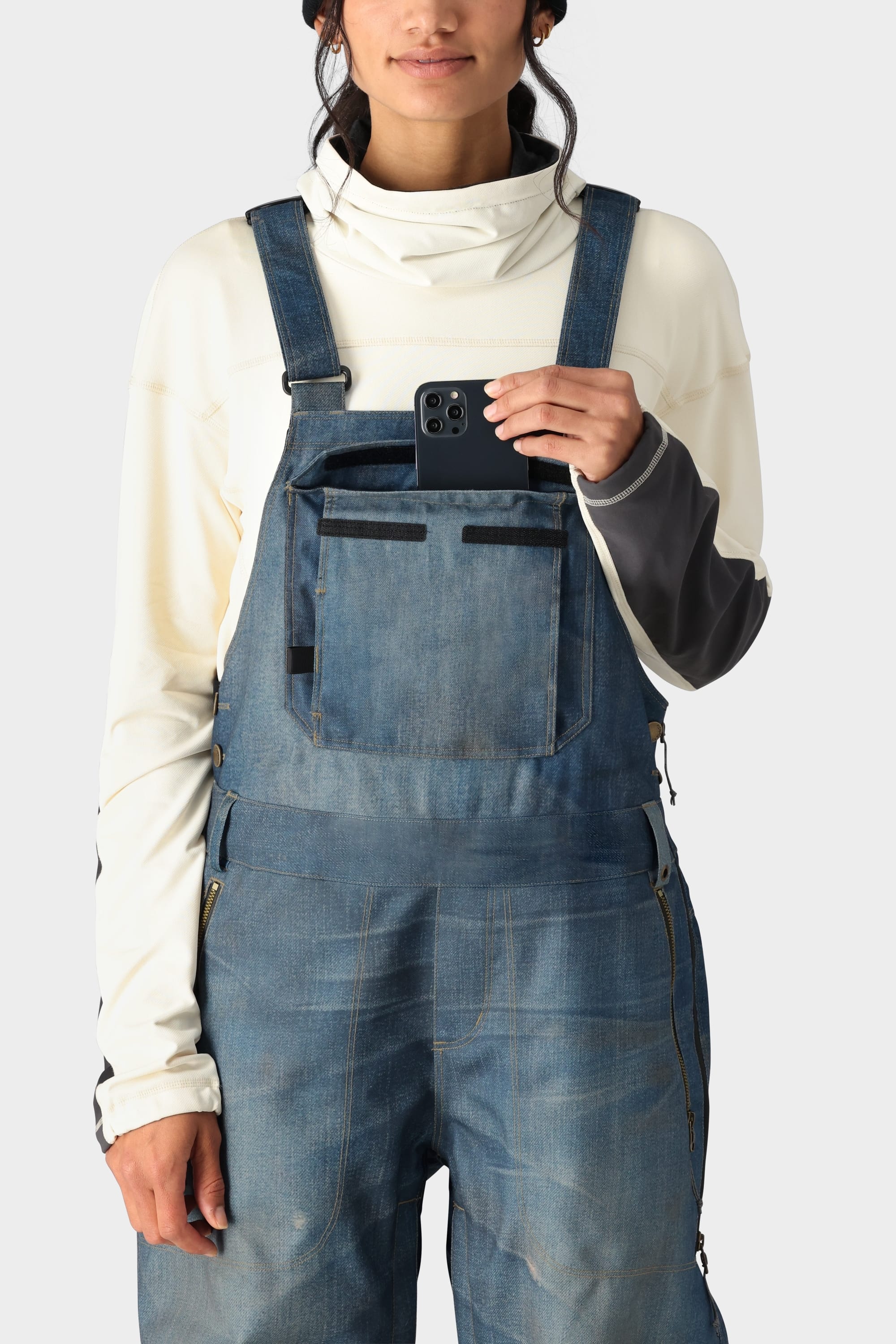 A person wearing the 686 Womens Black Magic 20K Bib by 686 is pocketing a smartphone in the front bib pocket. The background is plain light gray.