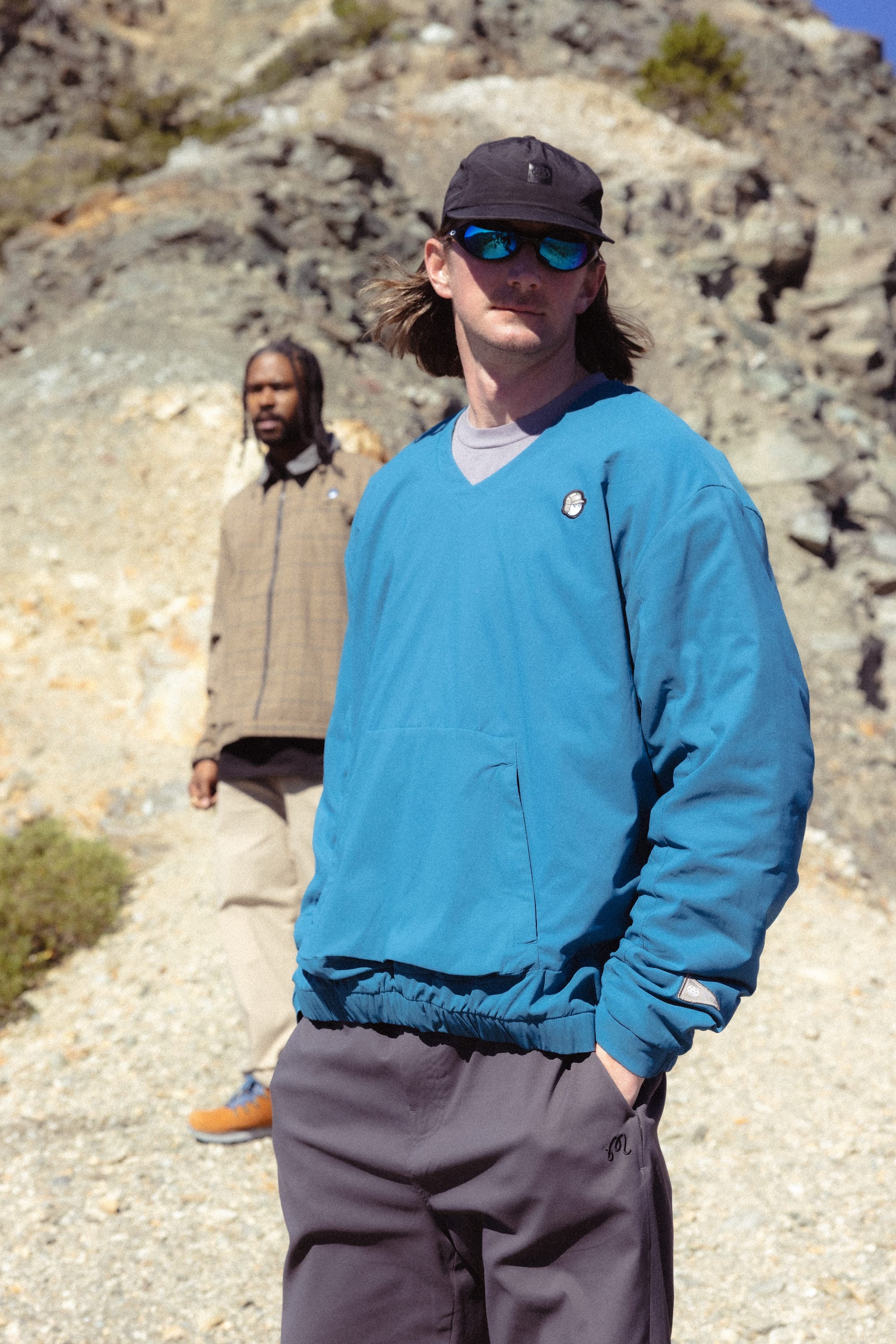 Two men stand outdoors on a rocky path; the man in front wears the 686 x Malbon Mens PrimaLoft drirelease® Merino-Lined Pullover Jacket, grey pants, a black cap, and sunglasses, while the man behind wears a beige jacket and light pants.