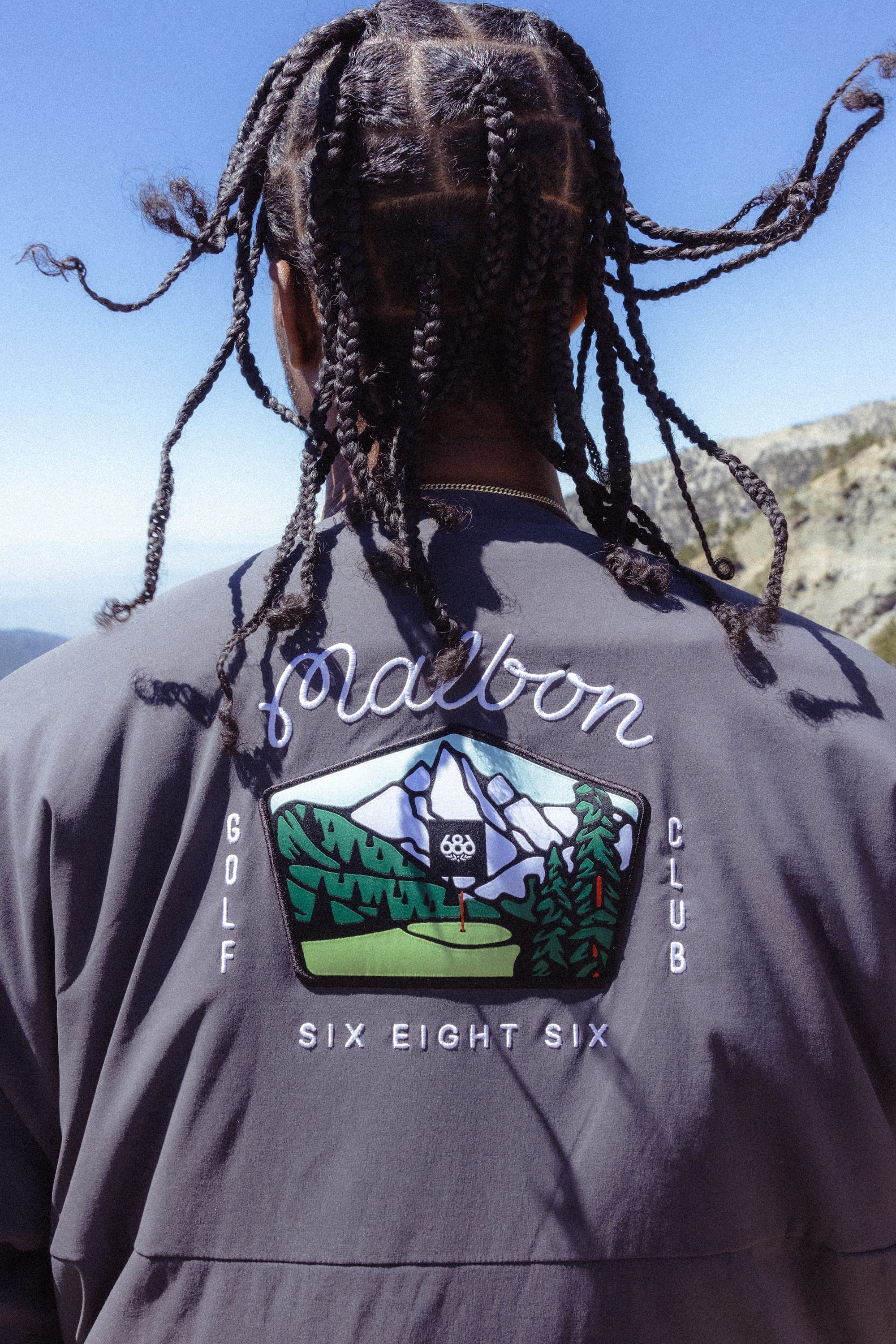 A person with braided hair stands outdoors facing away, wearing the 686 x Malbon Mens PrimaLoft drirelease® Merino-Lined Pullover Jacket featuring a colorful golf course patch labeled Malbon Golf Club and Six Eight Six.