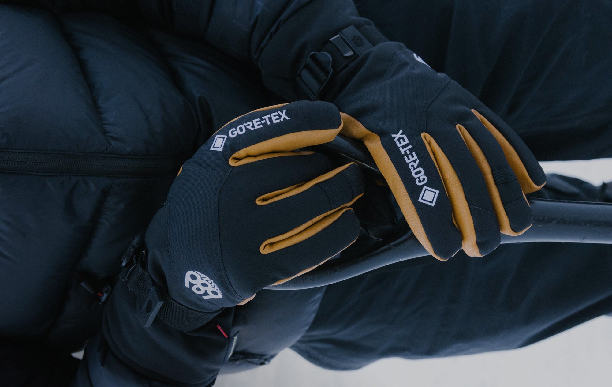 A close-up of hands wearing black and tan GORE-TEX gloves, resting on a black handlebar. The person is dressed in a dark, insulated jacket, suggesting cold weather or outdoor activity.