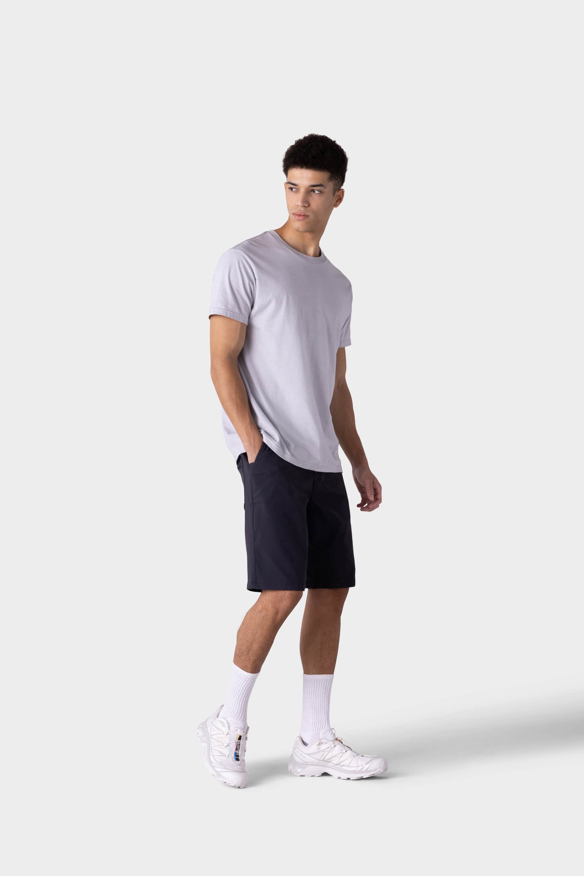 A young man stands against a plain light gray background wearing a light gray T-shirt, 686 Mens Everywhere Hybrid Short - Relaxed Fit by 686, white crew socks, and white sneakers. He looks slightly to his right with a relaxed posture.
