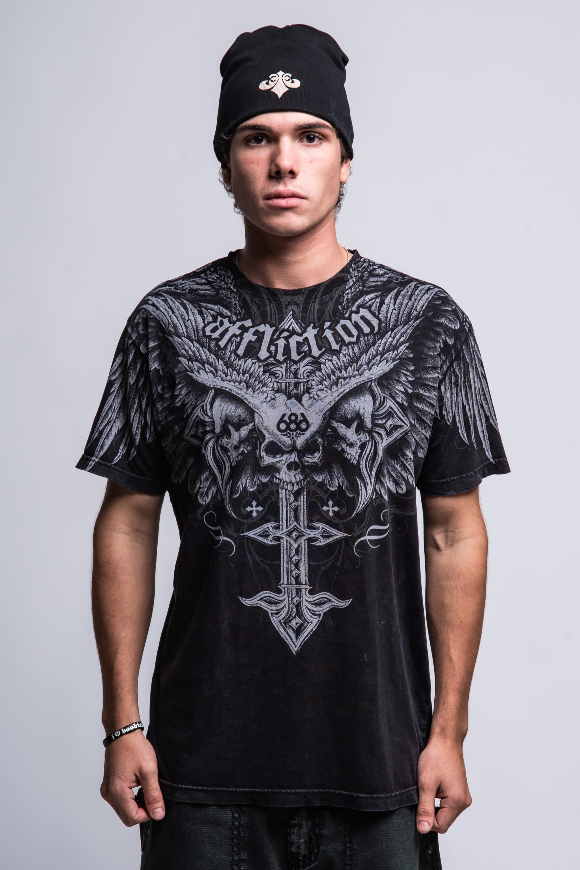 A young man in a black 686 Affliction Death Eyes SS Tee by 686 and a black beanie stands facing the camera against a plain light background. The tee features large, detailed eagle wings and Affliction text.