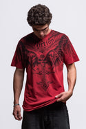 A person with curly dark hair looks down wearing the 686 Affliction Death Eyes SS Tee in red by 686, featuring black wings, a cross, and bold text, paired with black pants against a plain light background.