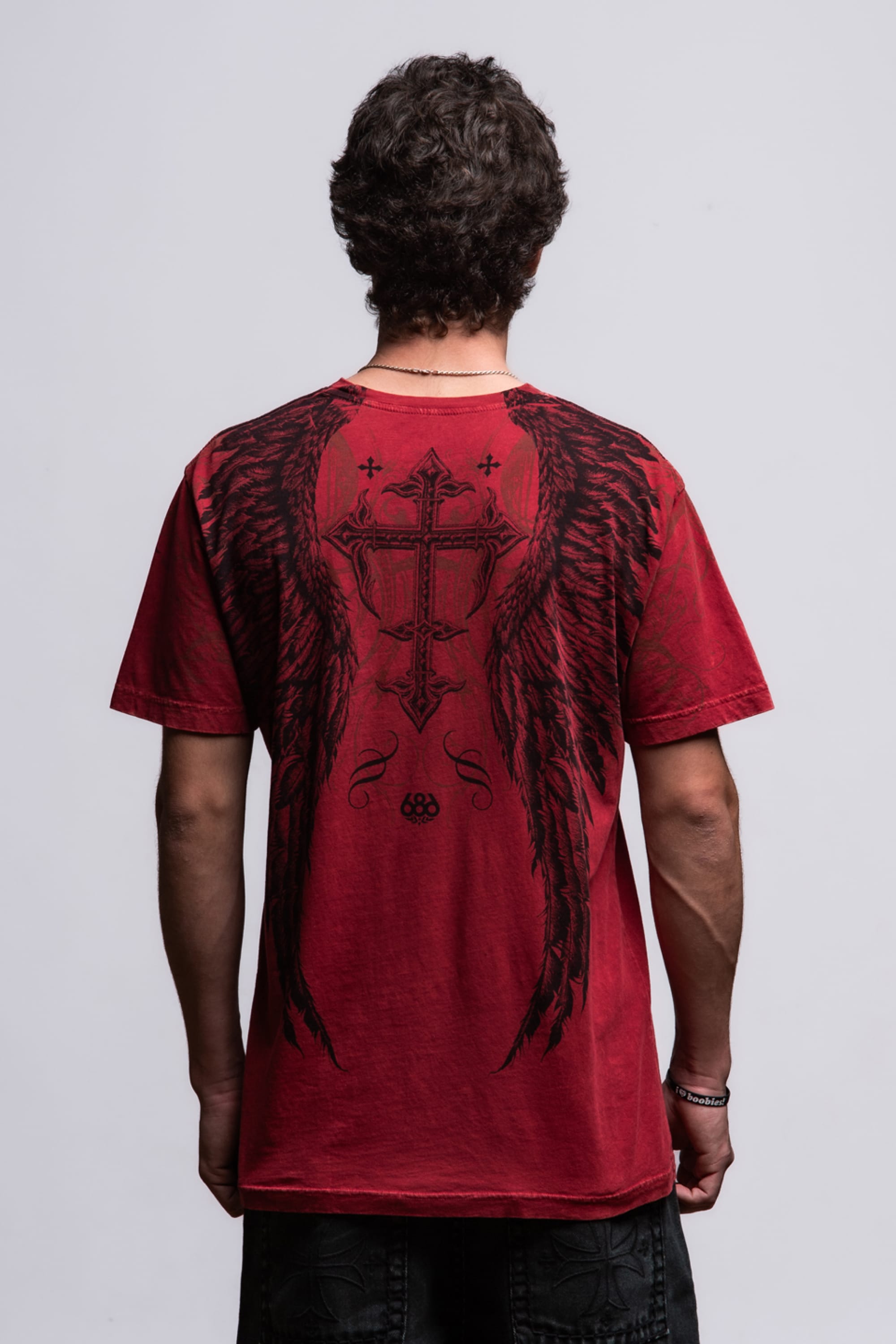 A person with short curly hair, seen from behind, wears the 686 Affliction Death Eyes SS Tee by 686 in red with large black wings and a cross on the back, standing before a plain light gray background.