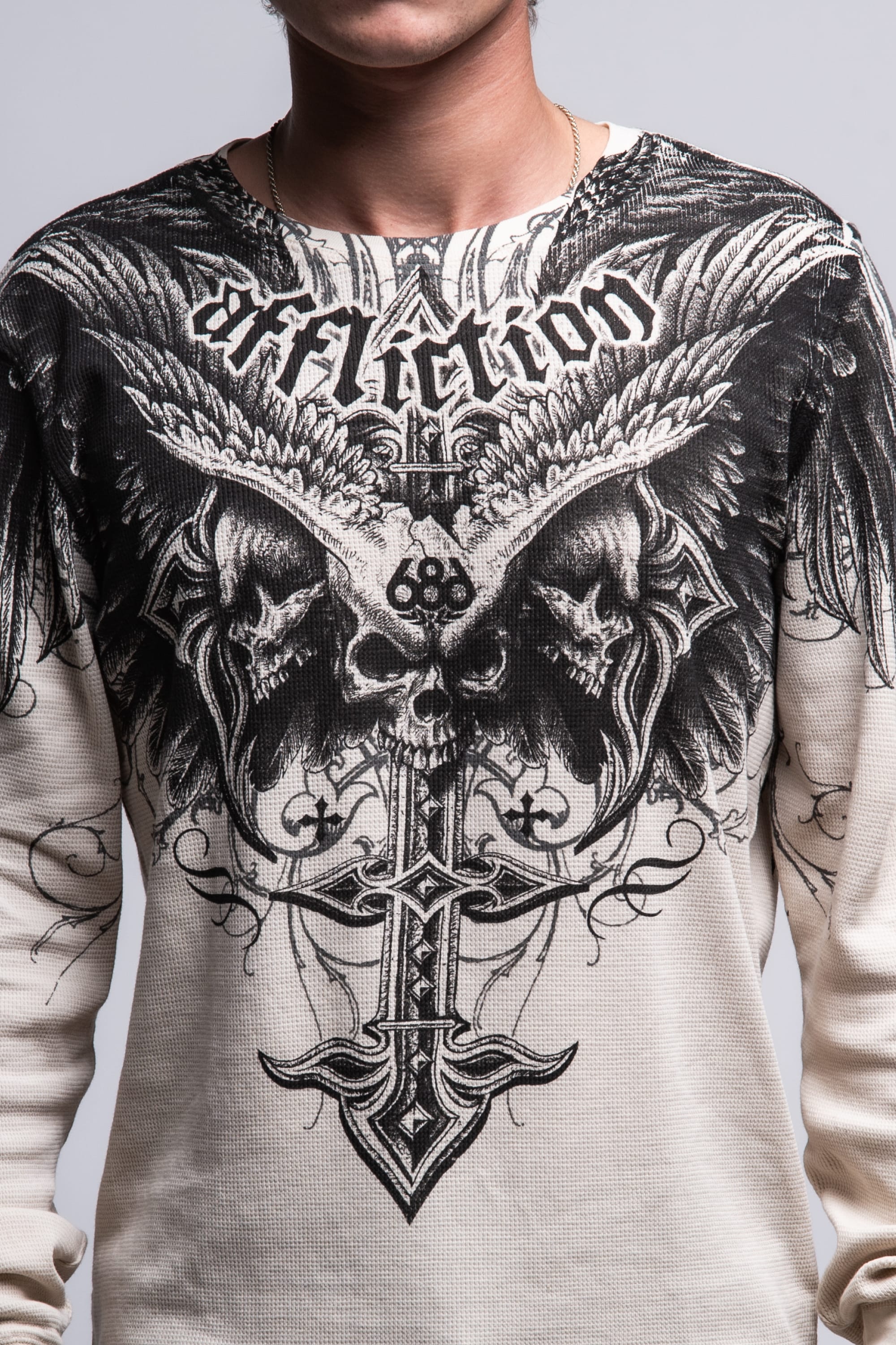 A person wears the 686 Affliction Death Eyes Thermal LS by 686—a light-colored, 100% cotton men’s tee with bold black graphics of skulls, wings, a cross, and “Affliction” in stylized text. Only their torso and part of neck are visible.