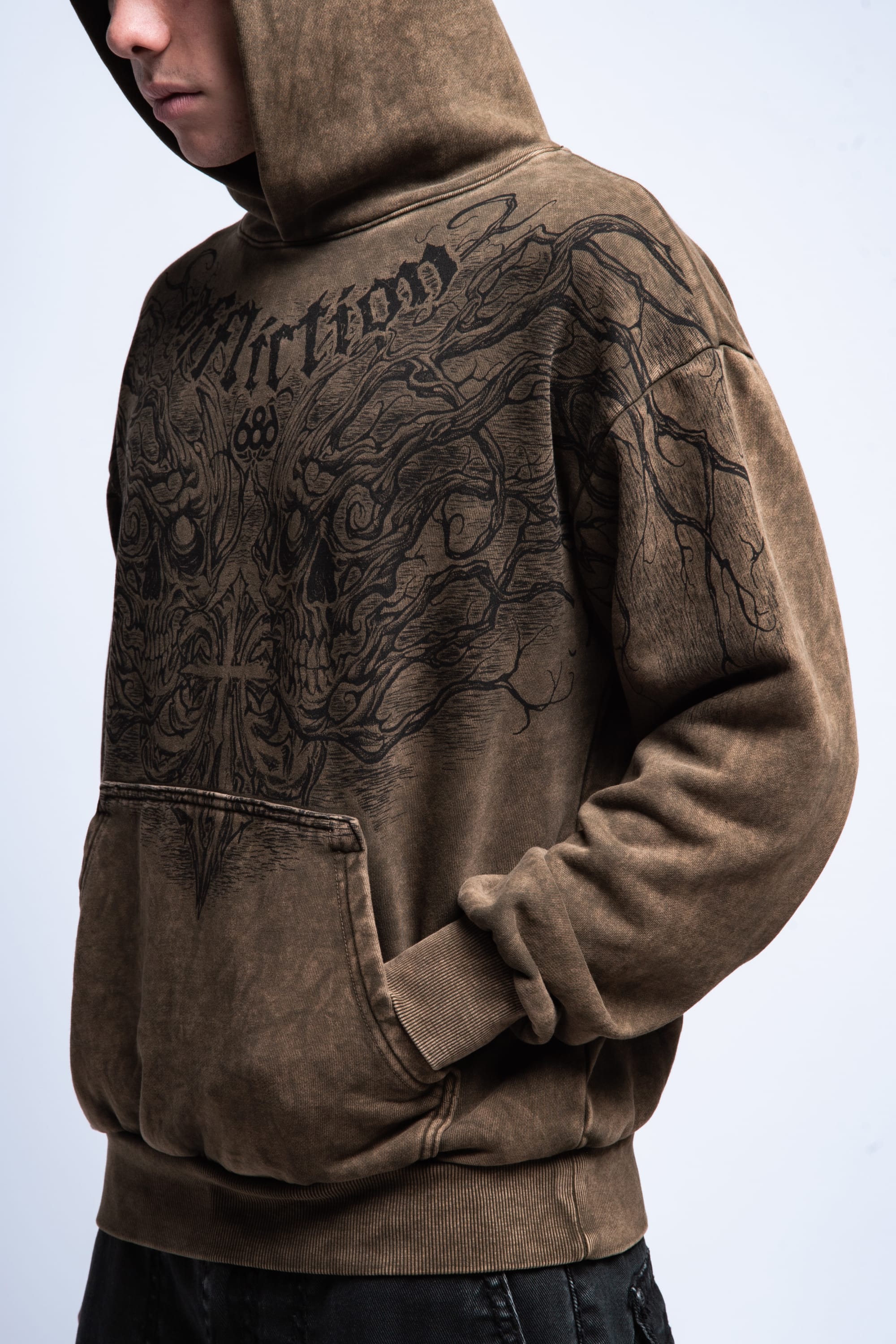 A person wearing the 686 Affliction Natural Rot Pullover Hoody by 686, featuring a dark, intricate front graphic and hood up, stands with hands in the kangaroo pocket against a plain light background.