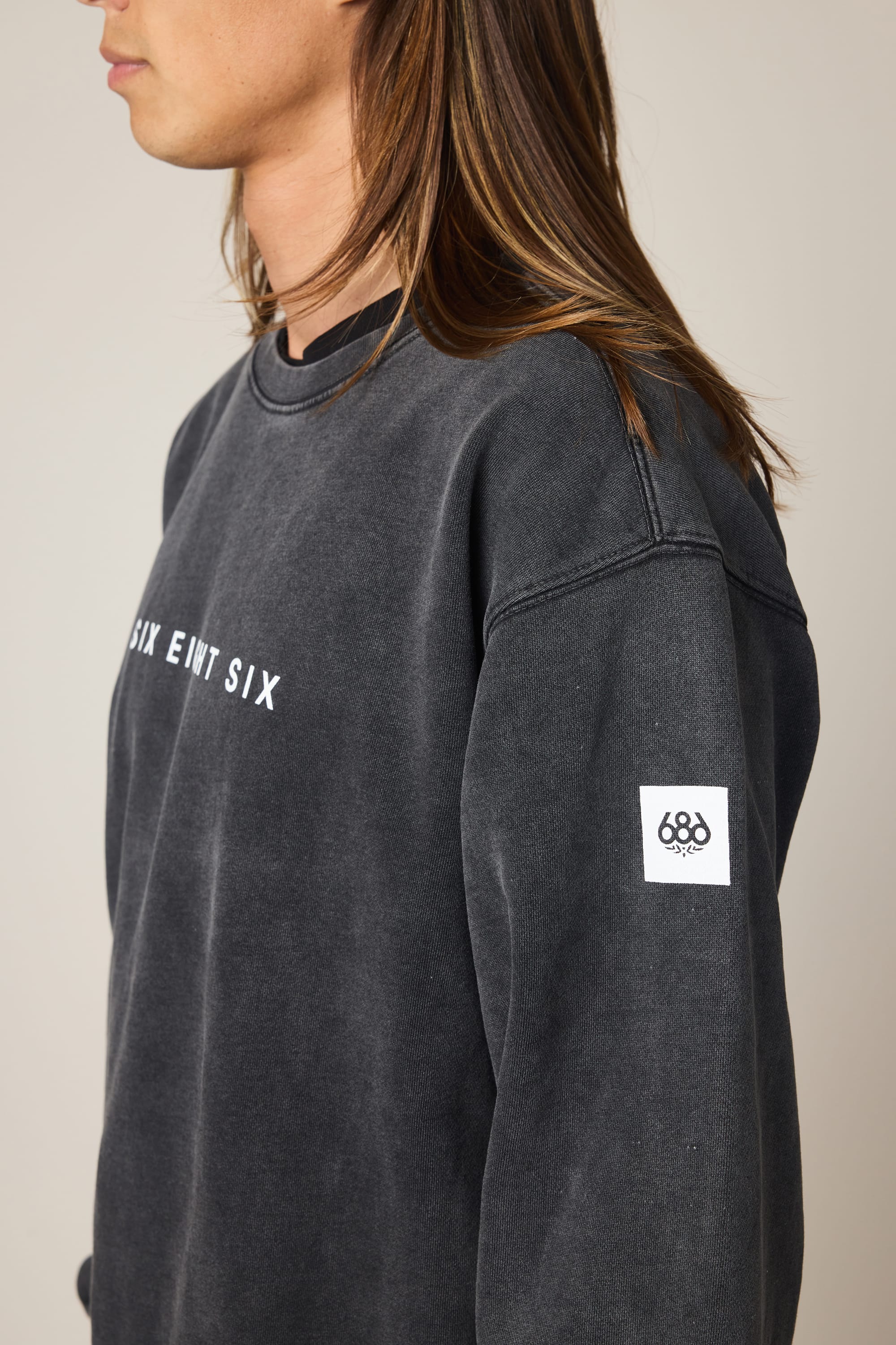 A person with long, light brown hair wears the 686 Men’s Mixed Relaxed LS Crewneck—a faded black 100% cotton sweatshirt with SIX EWT SIX text on the front and a white sleeve patch featuring the 686 logo, shown in side profile.