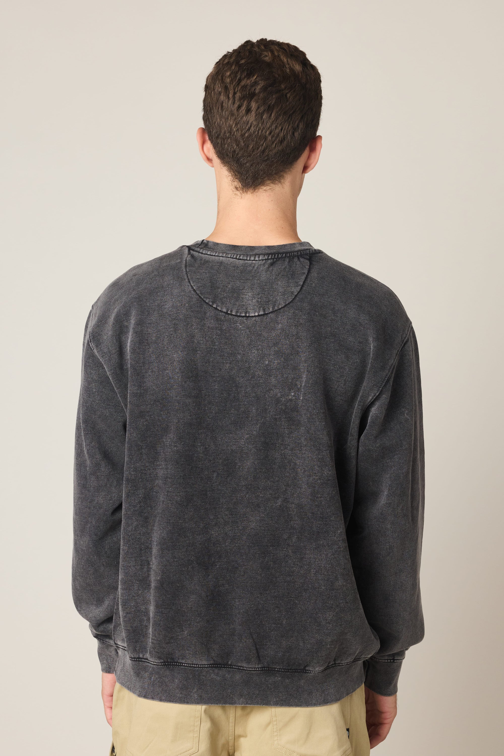 A man with short brown hair stands facing away from the camera, wearing the 686 Wreath Relaxed LS Crewneck by 686 in faded black and beige pants against a plain, light background.