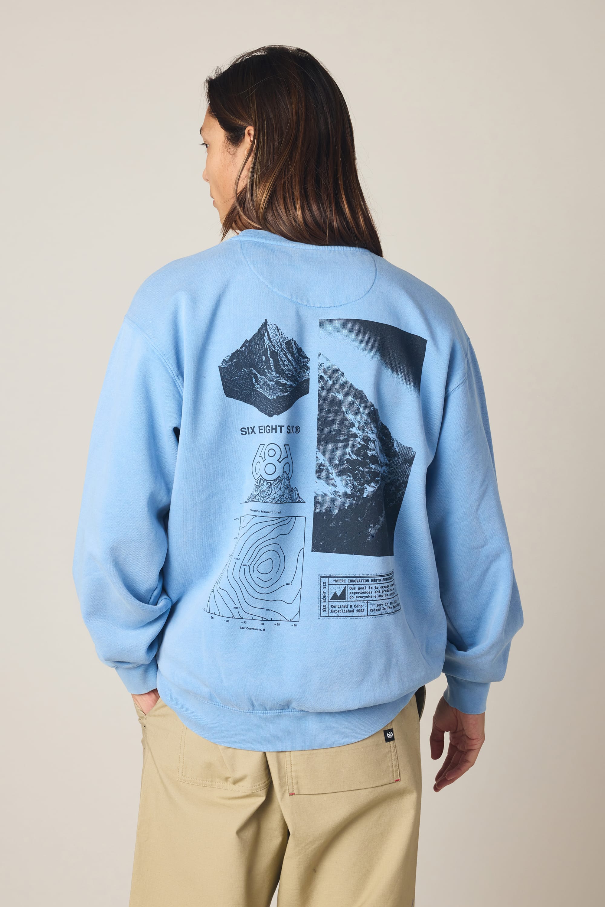 A person with long hair wears the 686 Men's Mixed Relaxed LS Crewneck in light blue, featuring black mountain graphics and topographic lines on the back, paired with beige pants against a plain background.