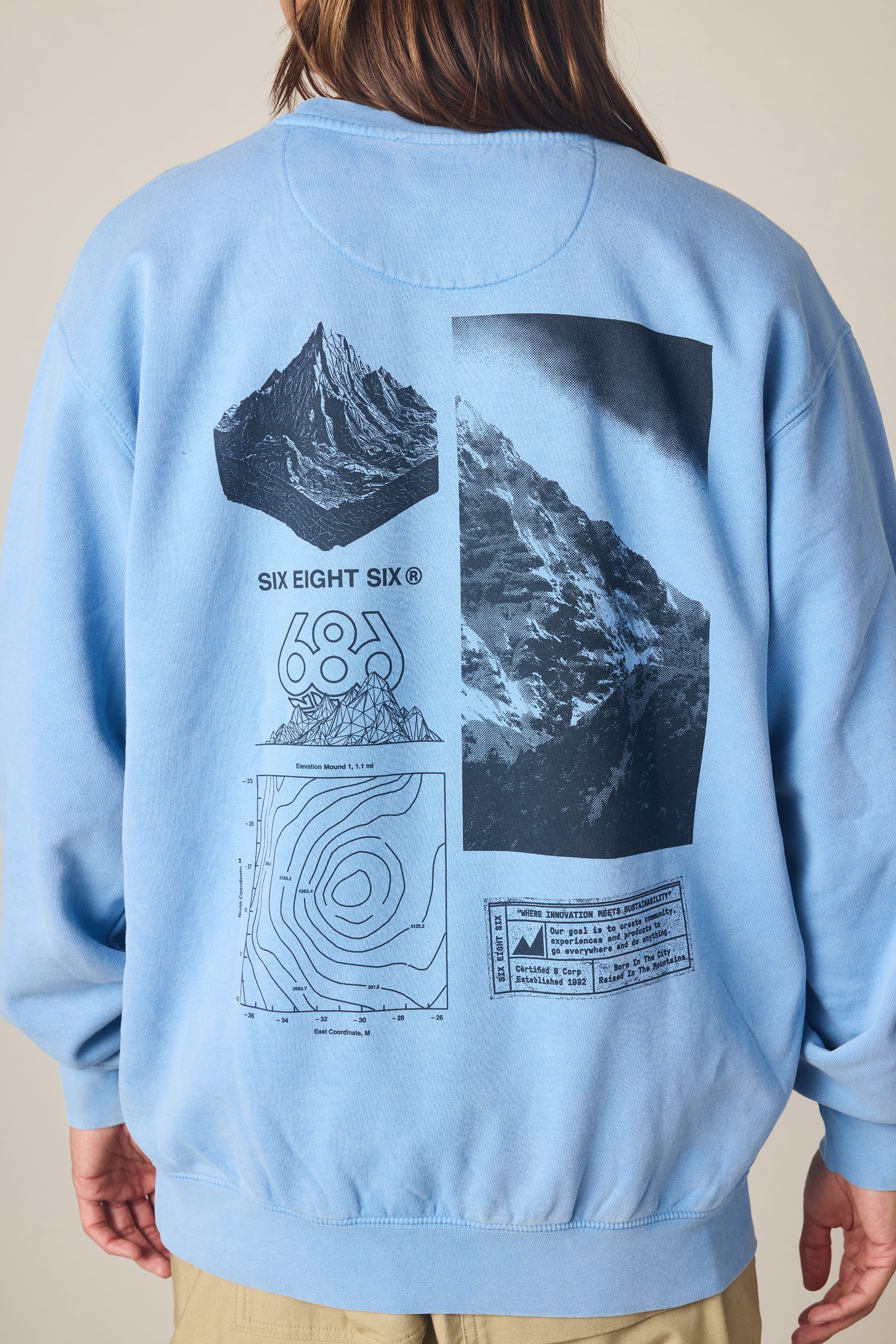 A person is standing facing away from the camera, wearing the 686 Men's Mixed Relaxed LS Crewneck in light blue. The 100% cotton sweatshirt features black graphics on the back, including mountain imagery, a contour map, various symbols, and small text.