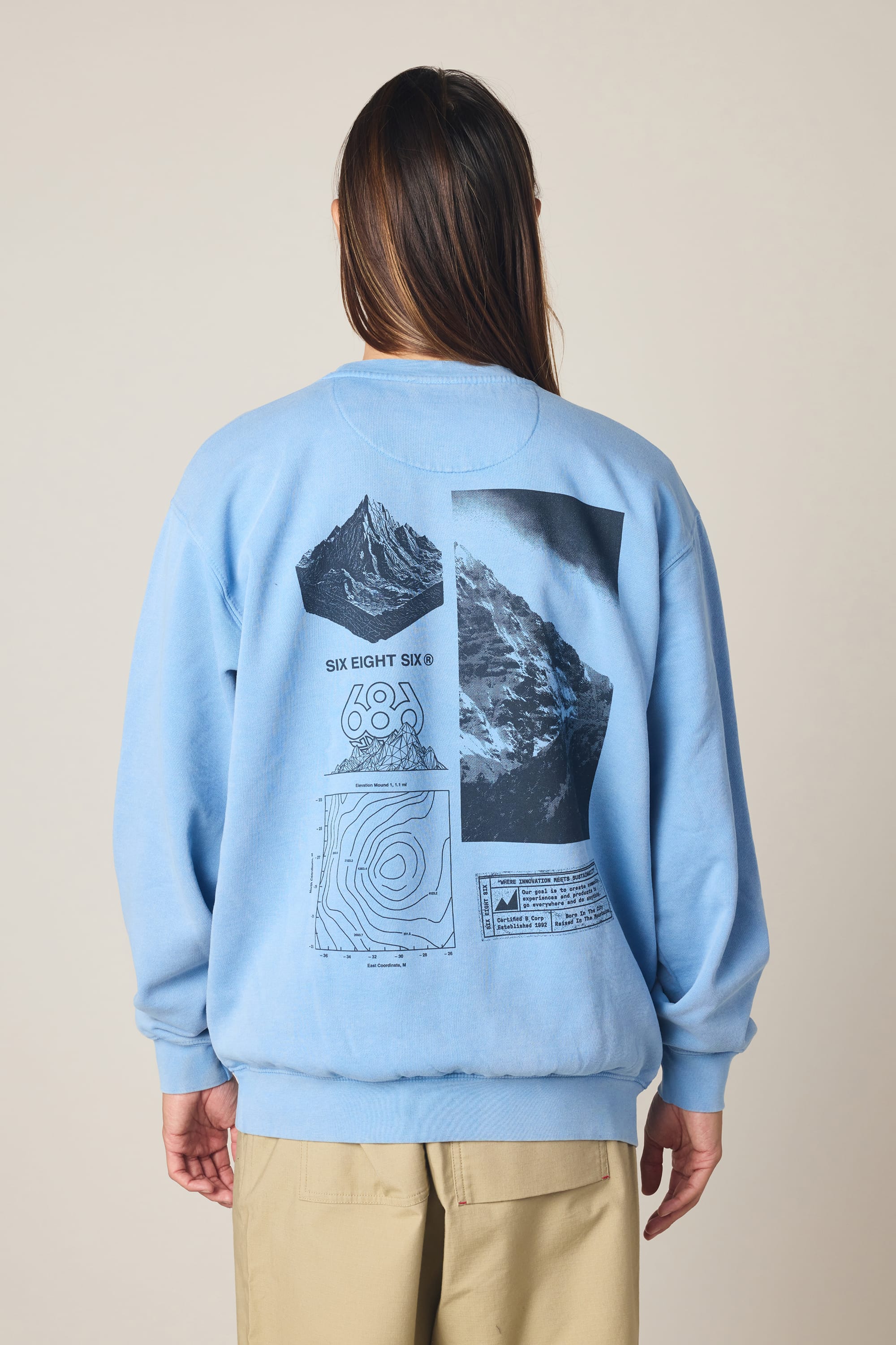 A person with long brown hair faces away from the camera, wearing a light blue 686 Men's Mixed Relaxed LS Crewneck by 686. The sweatshirt features black mountain graphics, a topographic map, and text on the back.