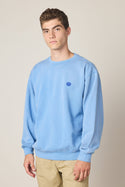 A young man with short brown hair wears the 686 Wreath Relaxed LS Crewneck by 686 with a small blue logo on the chest and beige pants, standing against a plain light background, looking at the camera with a neutral expression.