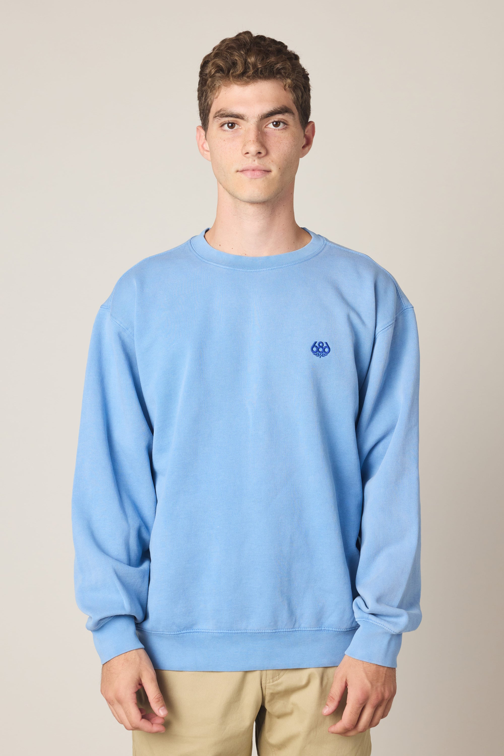 A young man with short curly hair wears the 686 Wreath Relaxed LS Crewneck—a light blue 100% cotton sweatshirt with a small embroidered logo on the chest—paired with beige pants, standing against a plain light background.