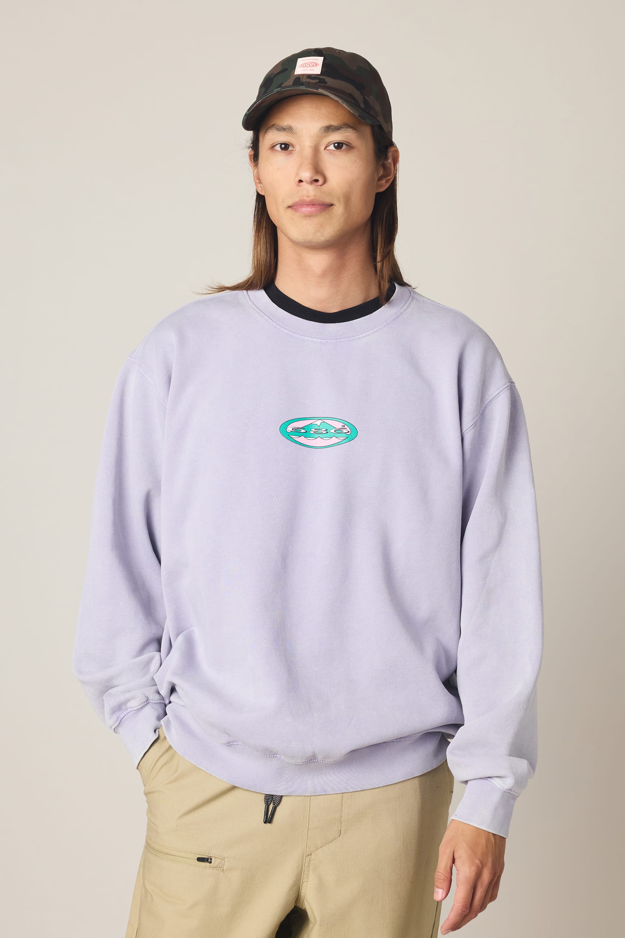 A person wearing the 686 Men's Mixed Relaxed LS Crewneck in lavender, featuring a small mountain logo, layered over a classic 100% cotton t-shirt, paired with beige pants and a black cap, stands against a neutral background and looks at the camera with a relaxed expression.