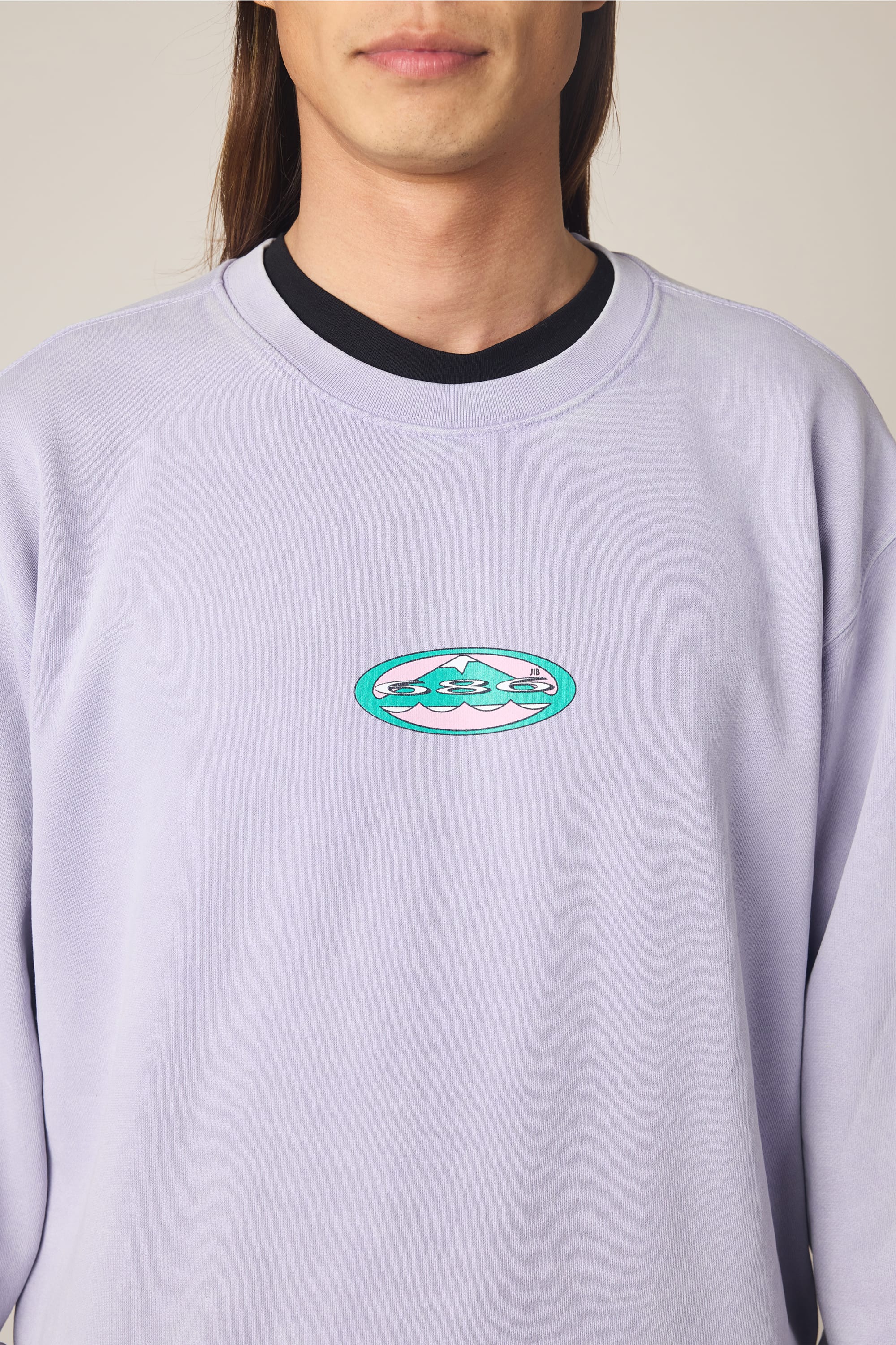 A person wears the 686 Men's Mixed Relaxed LS Crewneck in light purple, made of 100% cotton, featuring a small oval turquoise and white wavy logo on the chest. Their face is partially out of frame. Brand: 686.