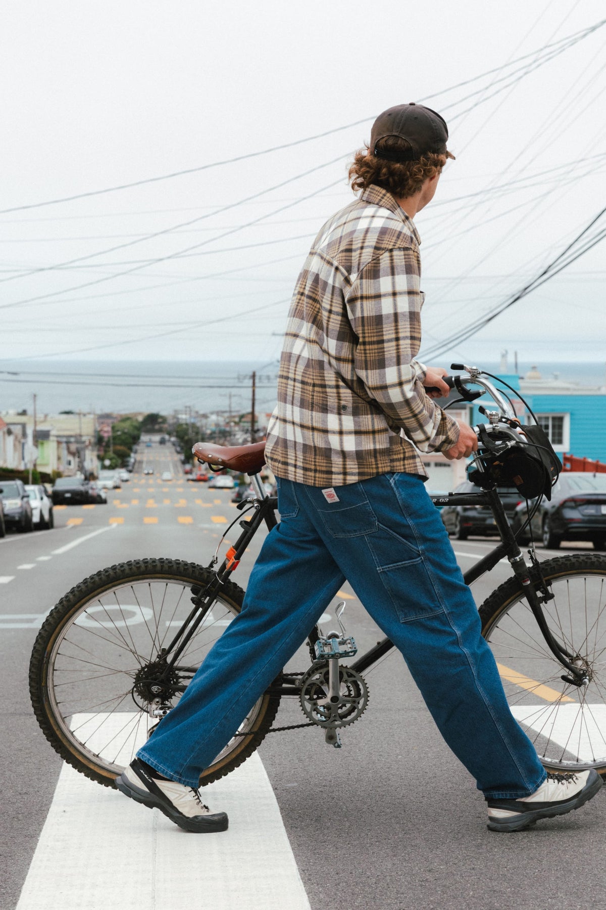 A man in a plaid shirt and 686 Men’s Unwork® Hammerhead Denim Pant walks a bicycle across a city street crosswalk.