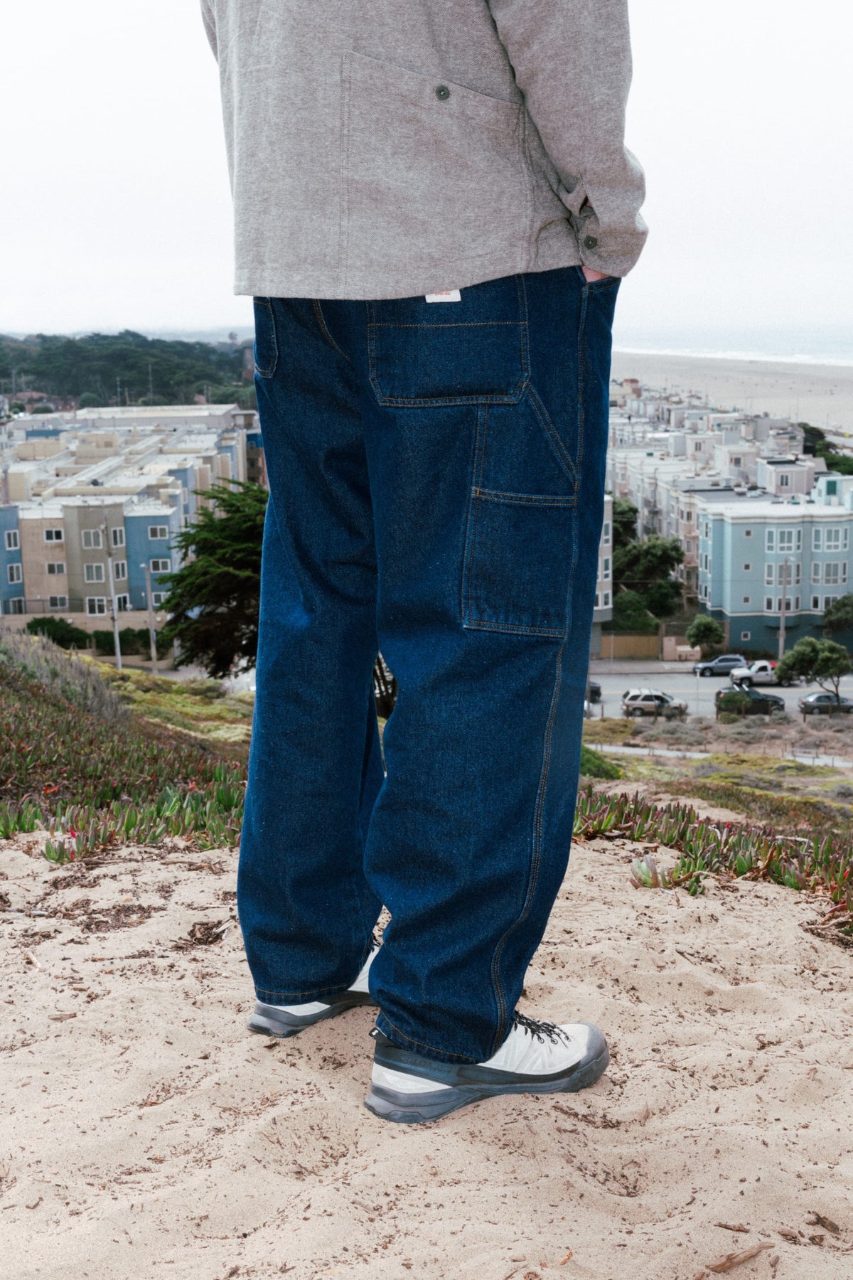 A person wearing baggy dark blue 686 Men’s Unwork® Hammerhead Denim Pant utility work jeans stands on a sandy hill overlooking a town and the ocean.