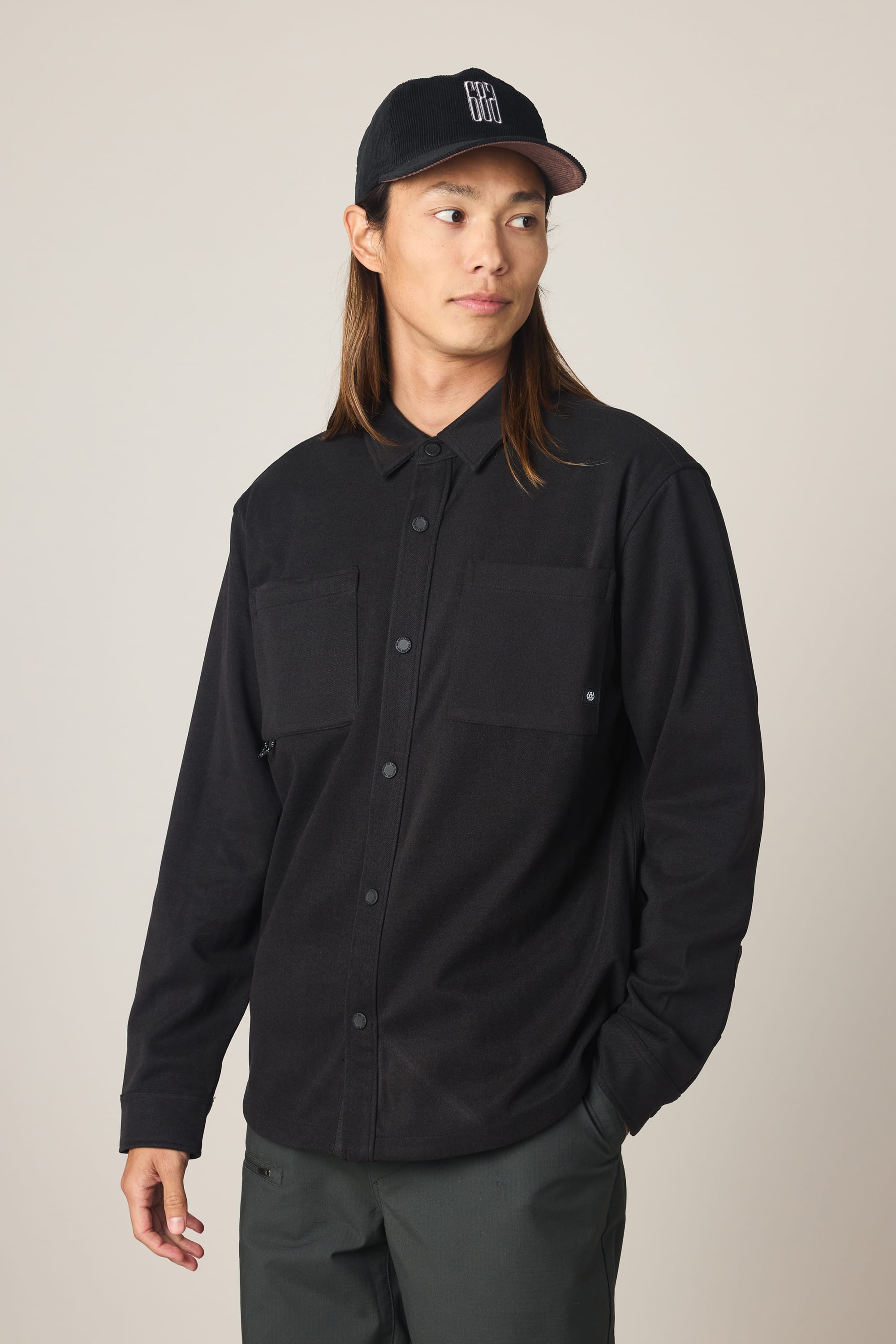A person with long brown hair wears the 686 Men's Whisper Knit Flannel and a black baseball cap, paired with dark pants, standing against a plain light background and looking to the side with one hand in their pocket.