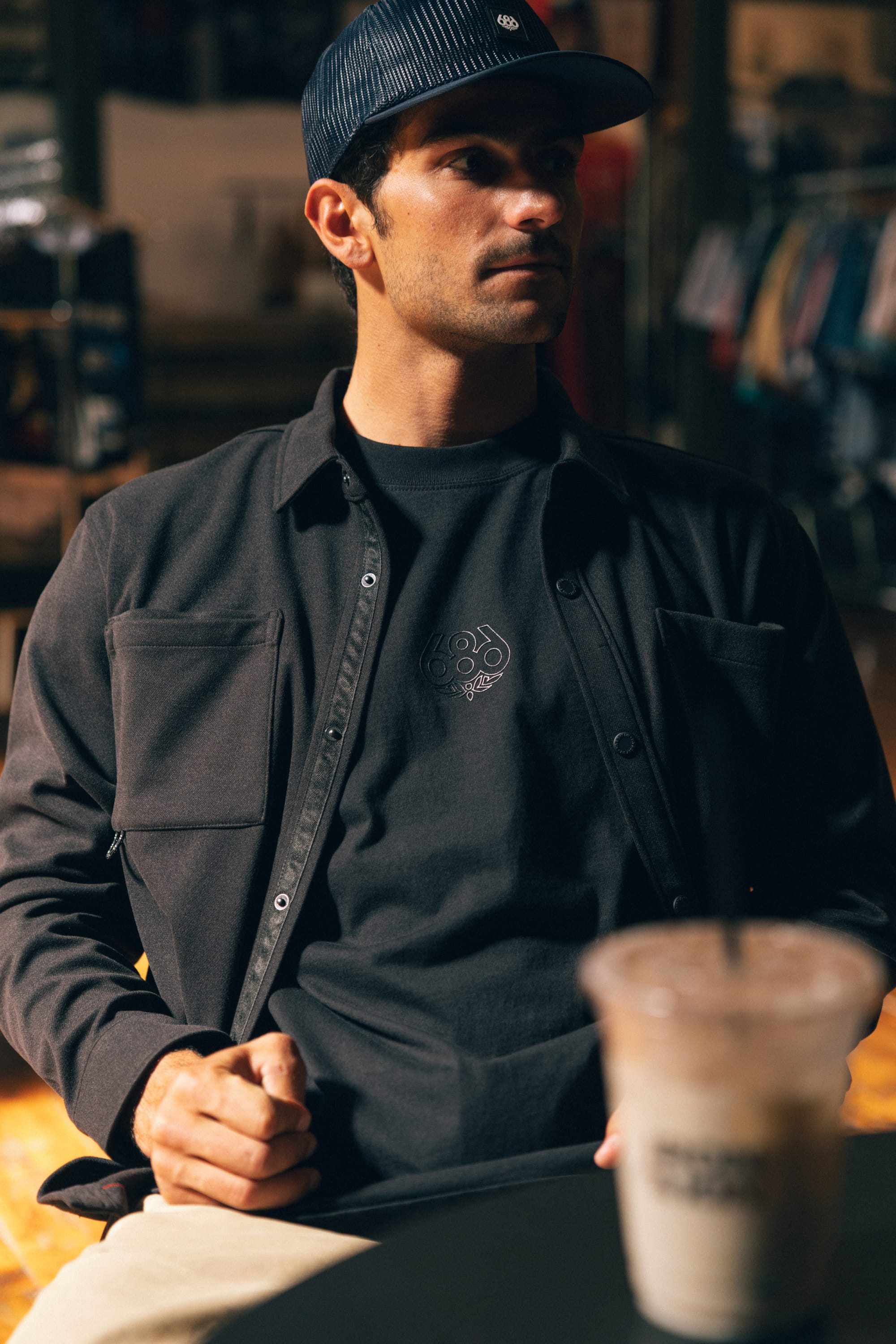 A man wearing the 686 Men's Whisper Knit™ Flannel by 686 and a striped cap sits indoors at a table with a blurry iced coffee in the foreground. Clothing racks are visible in the background, and his stylish polyester flannel adds a touch of modern style to his relaxed look.