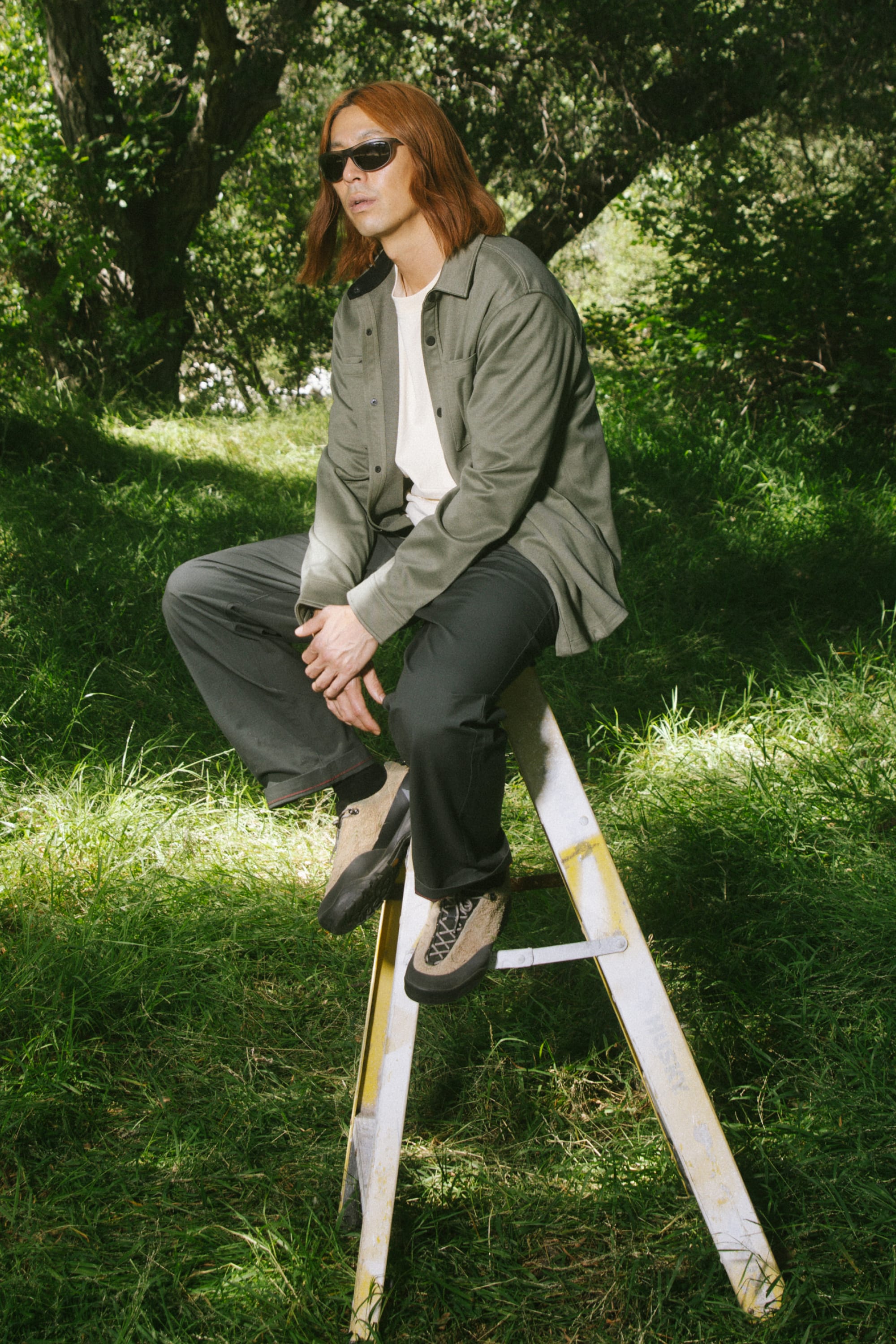 A person with long, reddish hair wearing sunglasses, a green jacket, the 686 Men's Whisper Knit™ Flannel from 686, and dark pants sits on a stepladder in a grassy, wooded area.