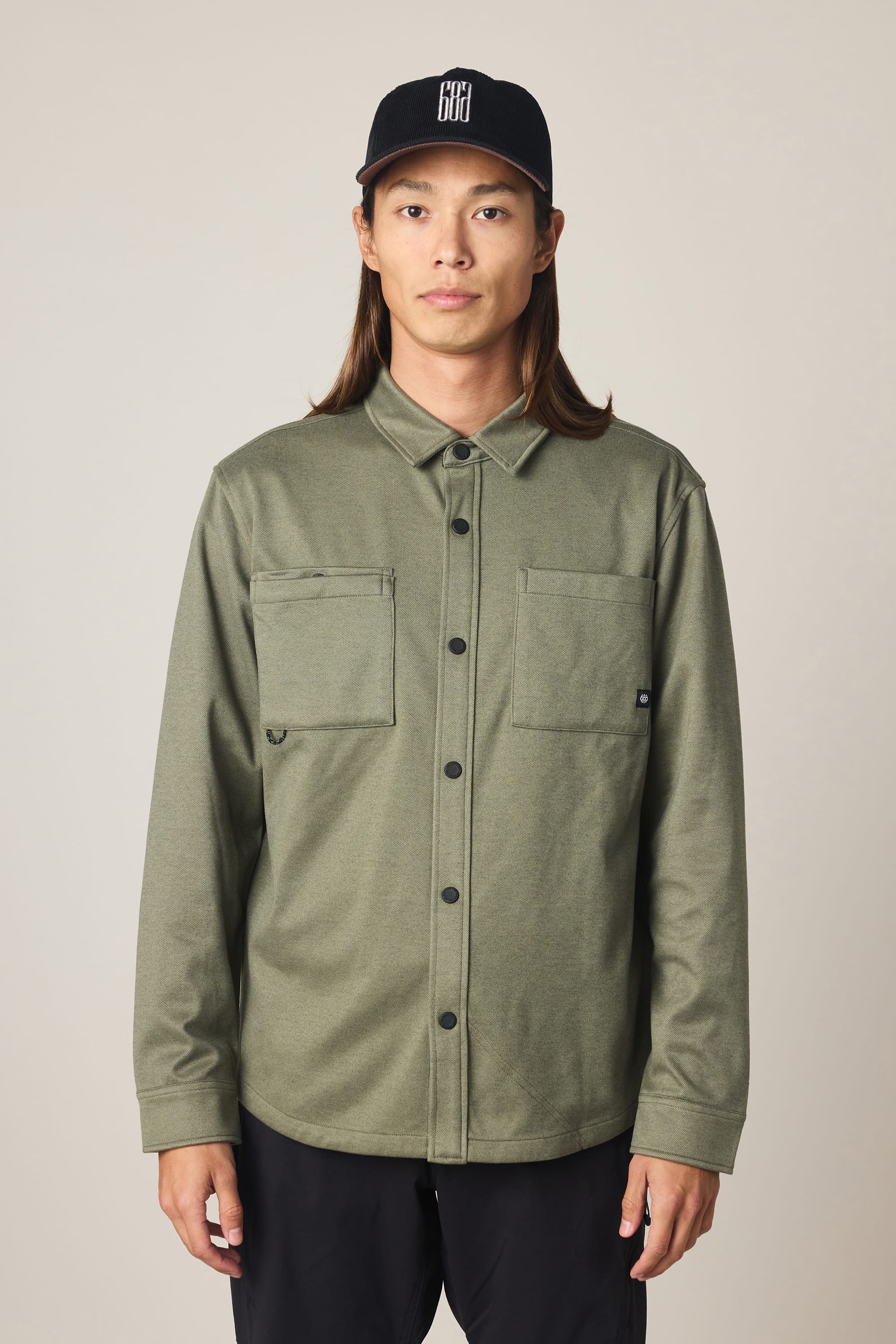 A person with long brown hair wears the 686 Men's Whisper Knit Flannel by 686—an olive green shirt with two chest pockets and black buttons—paired with black pants and a black baseball cap, standing against a neutral background.