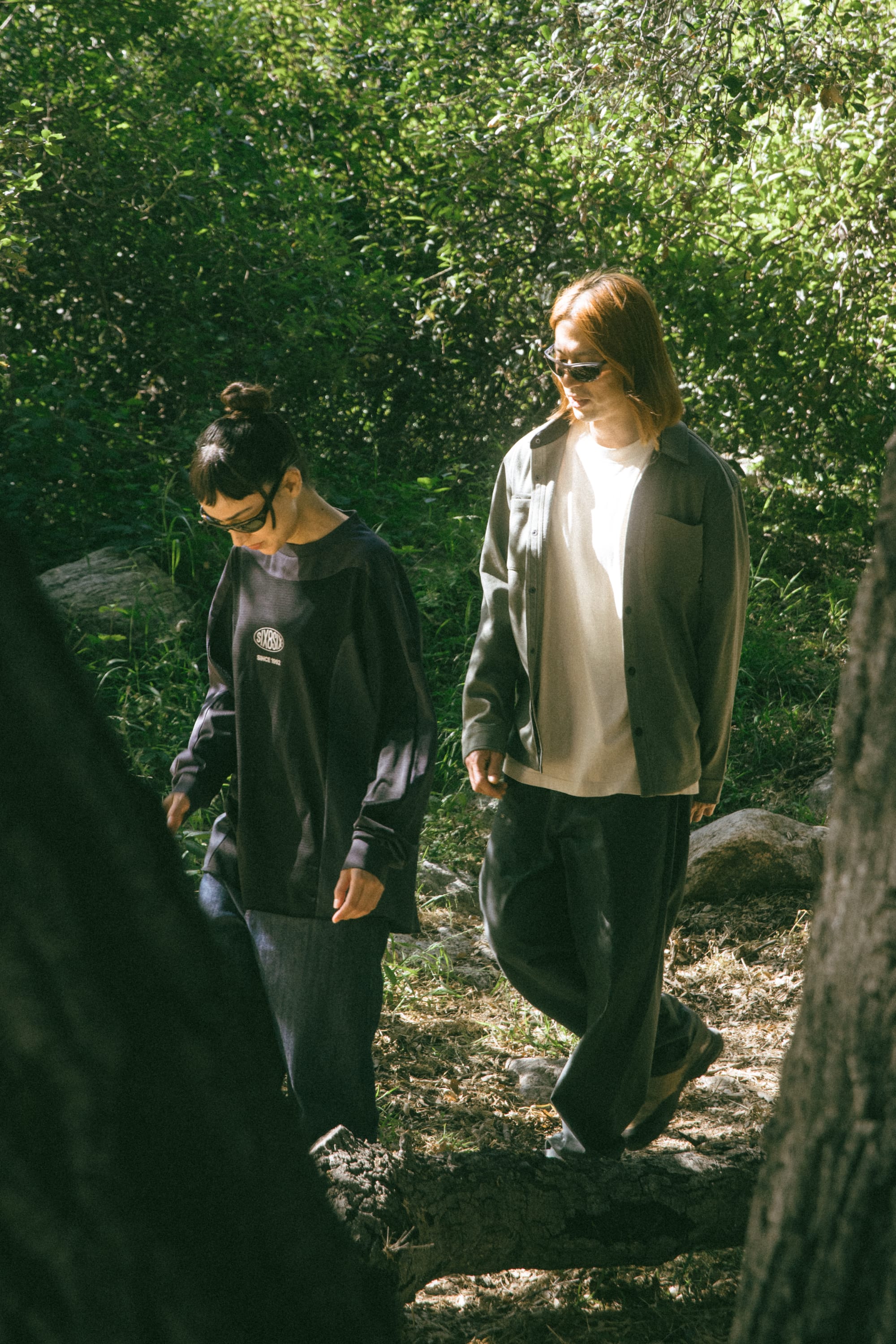 Two people wearing sunglasses and casual clothing, including a stylish 686 Men's Whisper Knit™ Flannel by 686 made from 100% polyester, walk together on a trail in a sunlit forest surrounded by green foliage and trees.