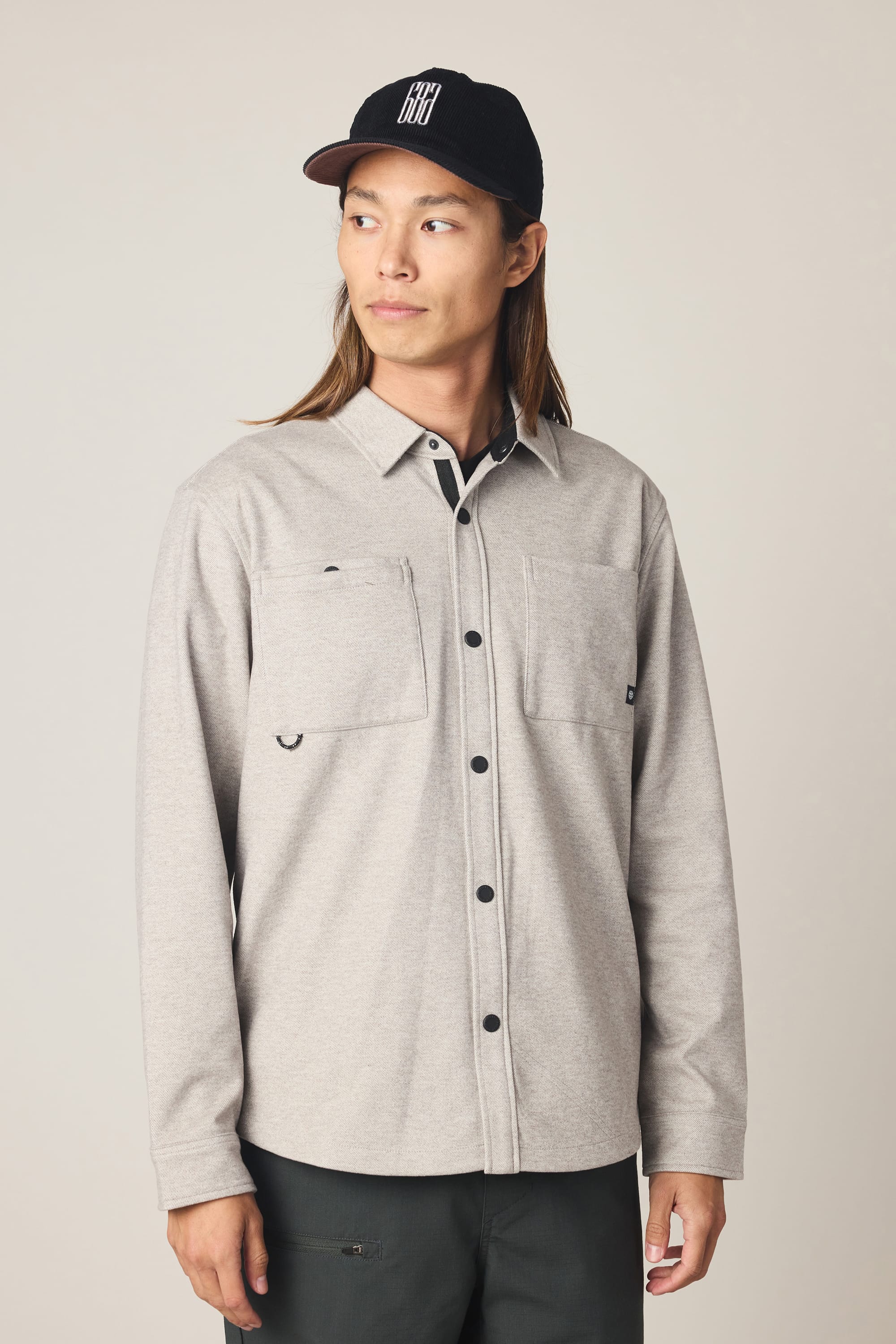 A person with long hair wears a black cap, dark pants, and a light gray 686 Men's Whisper Knit Flannel shirt with two chest pockets, standing against a plain background and looking to the side.