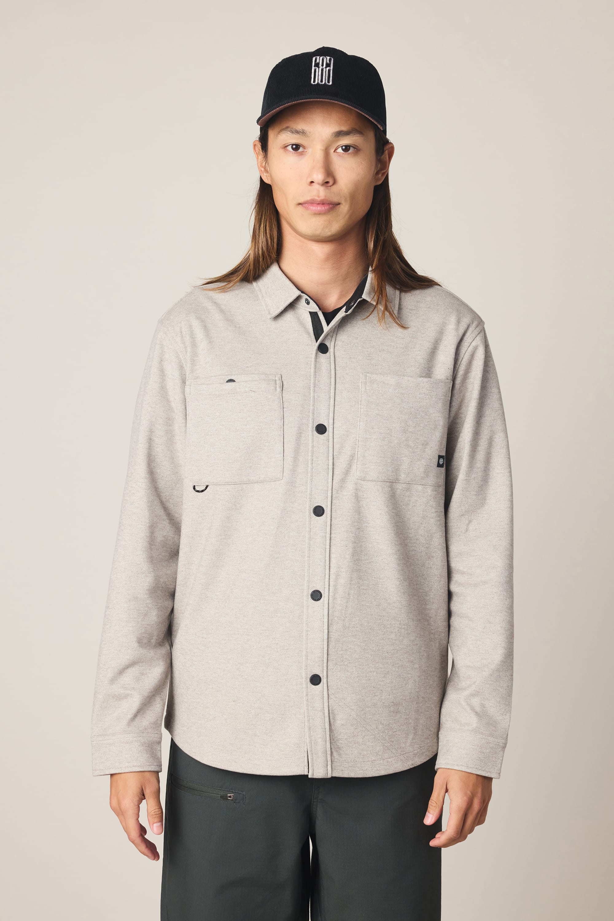 A person with long brown hair, wearing a black baseball cap, the 686 Men's Whisper Knit™ Flannel by 686—a light gray men's shirt made of 100% polyester with two chest pockets—and dark gray pants stands facing forward against a plain beige background.