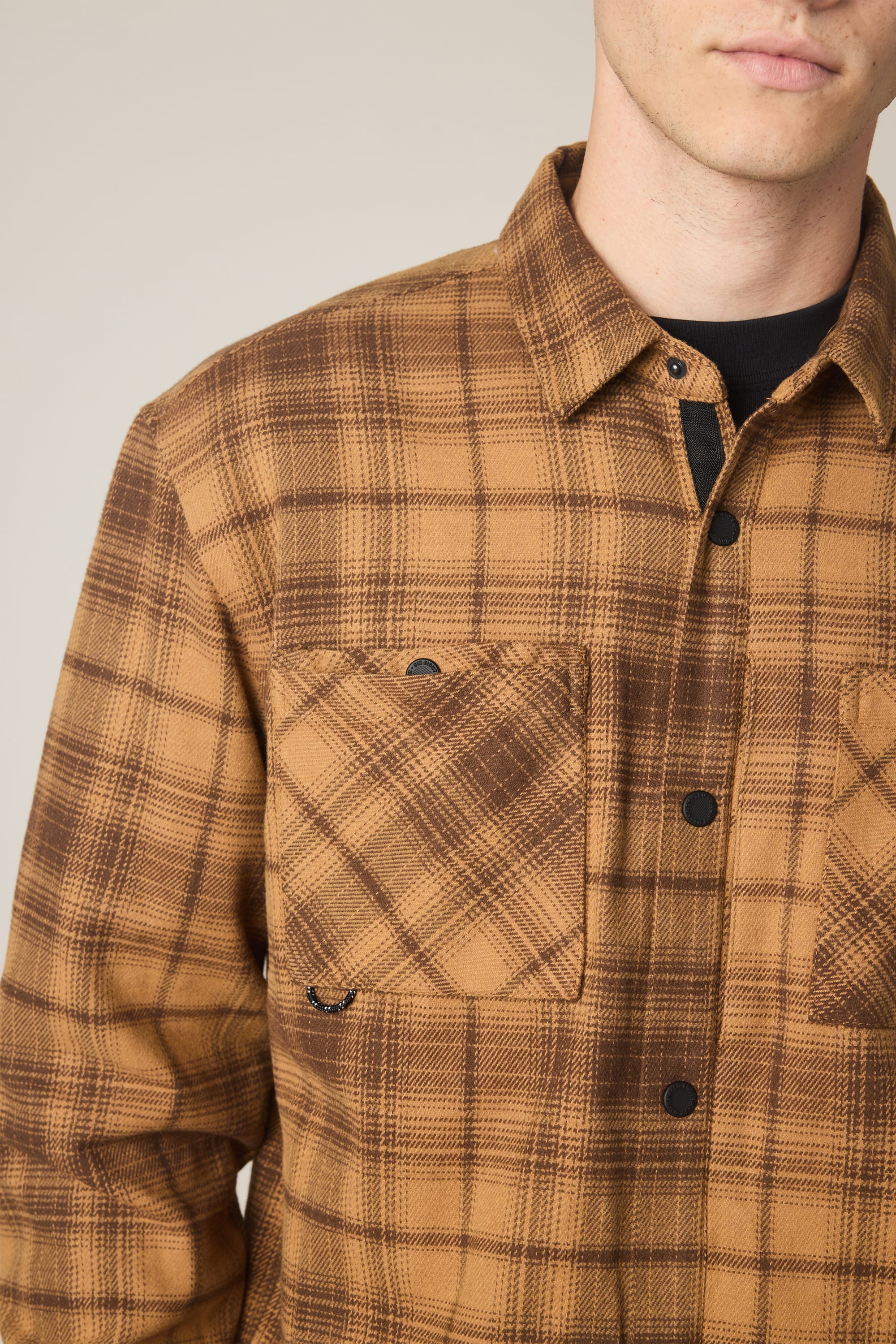 A close-up shows a person wearing the 686 Men's Heat Grid Flannel in brown/tan plaid, featuring two chest pockets. The photo is cropped at the shoulders and mouth, displaying only the lower face and upper torso.