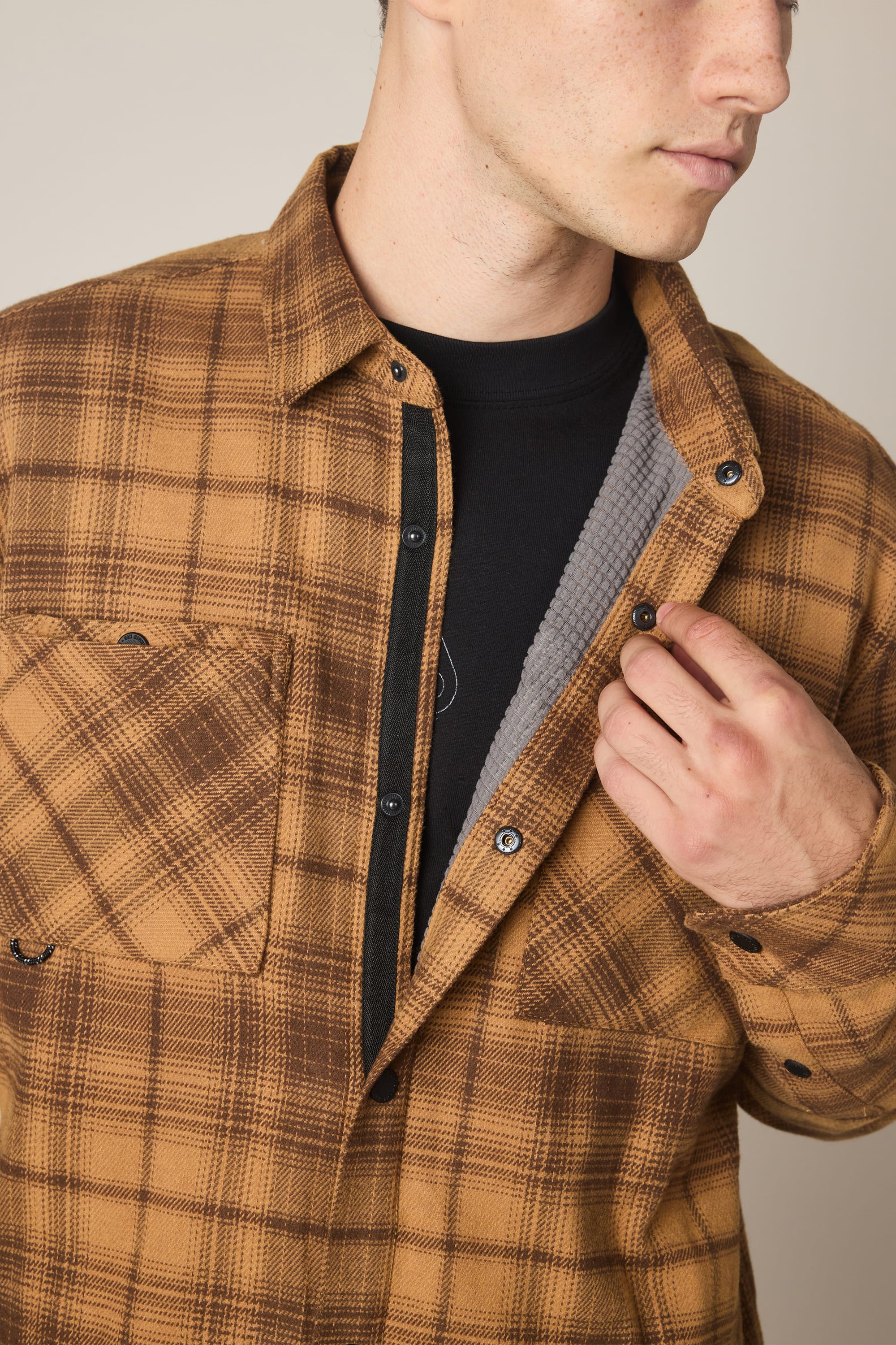 A person, partially out of frame, wears the 686 Men's Heat Grid Flannel by 686—brown plaid polyester over a black shirt—holding it open to show the gray textured lining inside.