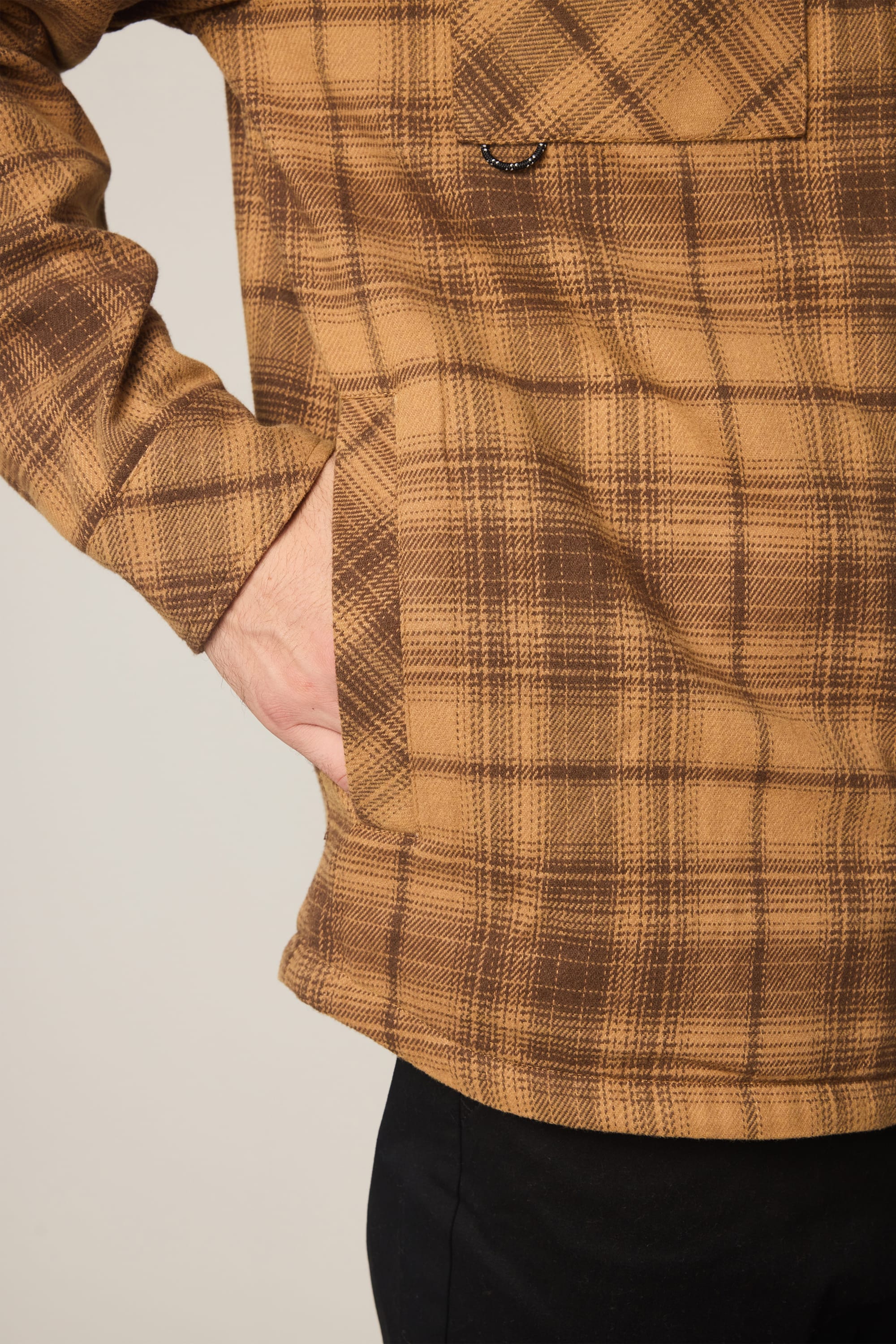 A person wearing the 686 Men's Heat Grid Flannel, a brown plaid polyester shirt by 686, has one hand in the front pocket. The lower part of the shirt and a black pant leg are shown against a plain background.