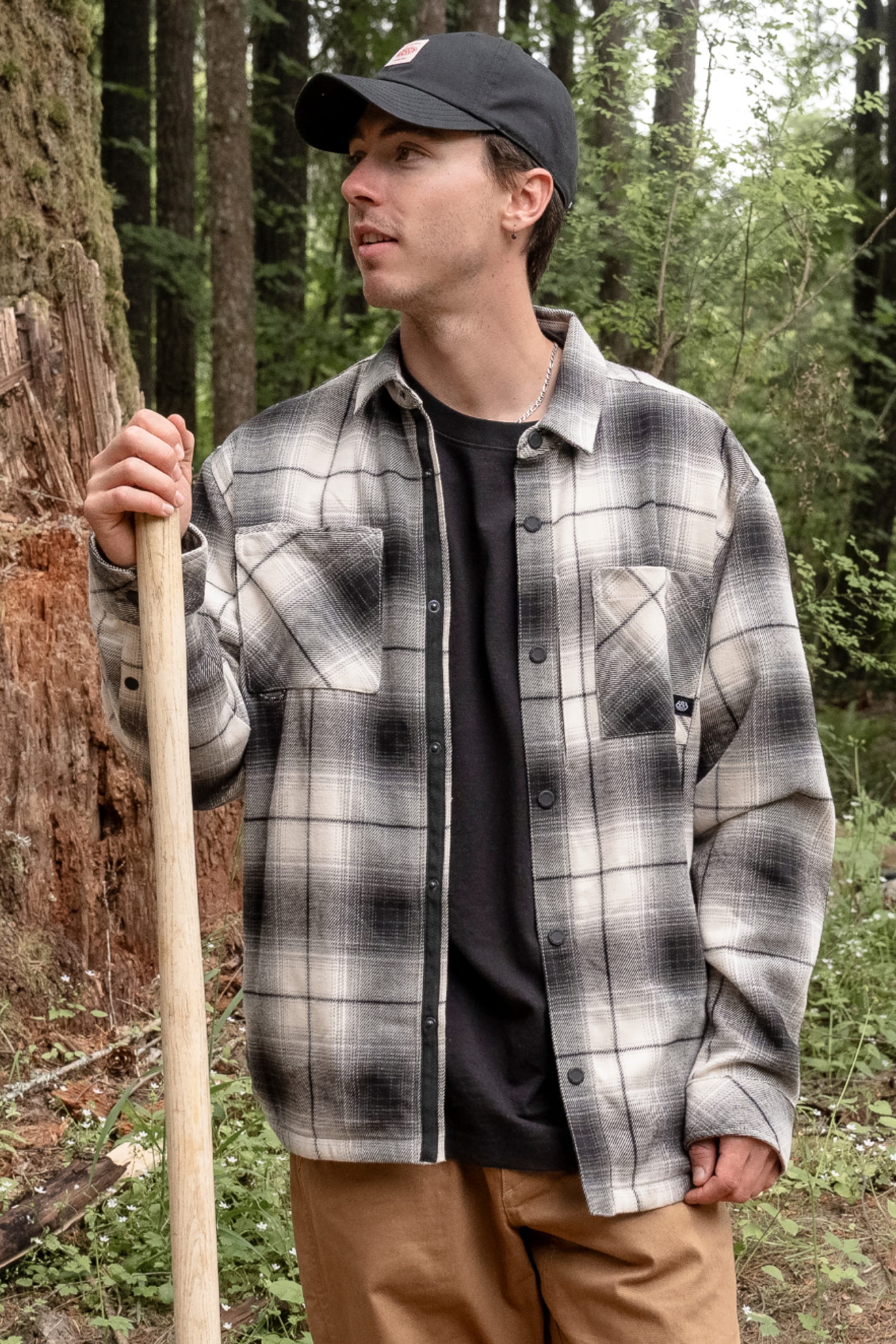 A man wearing the 686 Men's Heat Grid™ Flannel by 686, along with a black cap and tan pants, stands in a forest holding a wooden stick and looking to the side. He is surrounded by green trees and a fallen log.