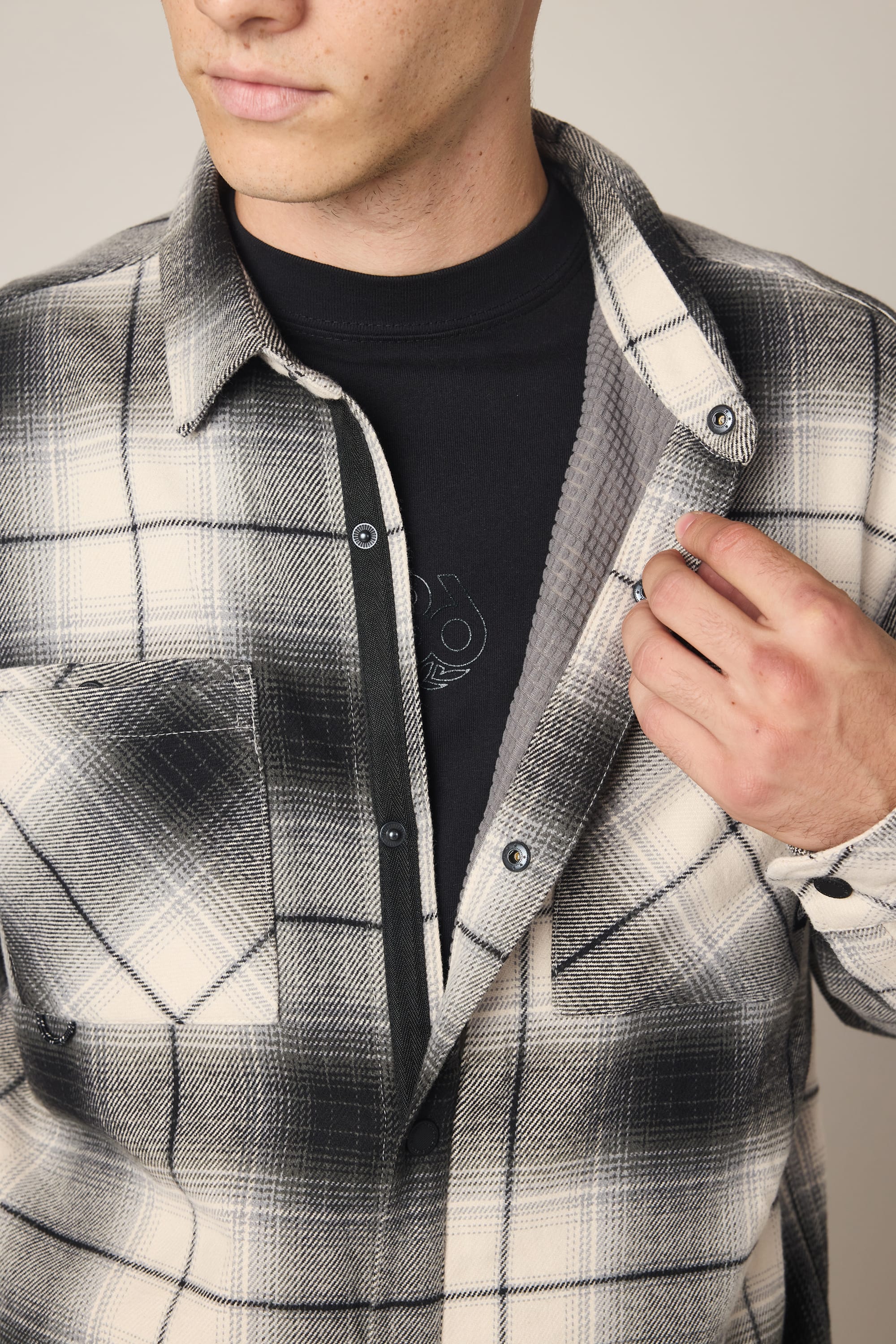 A person is shown unbuttoning a 686 Men's Heat Grid Flannel over a black T-shirt. The flannel features a black-and-white plaid pattern. The image is cropped at the shoulders and mid-torso.