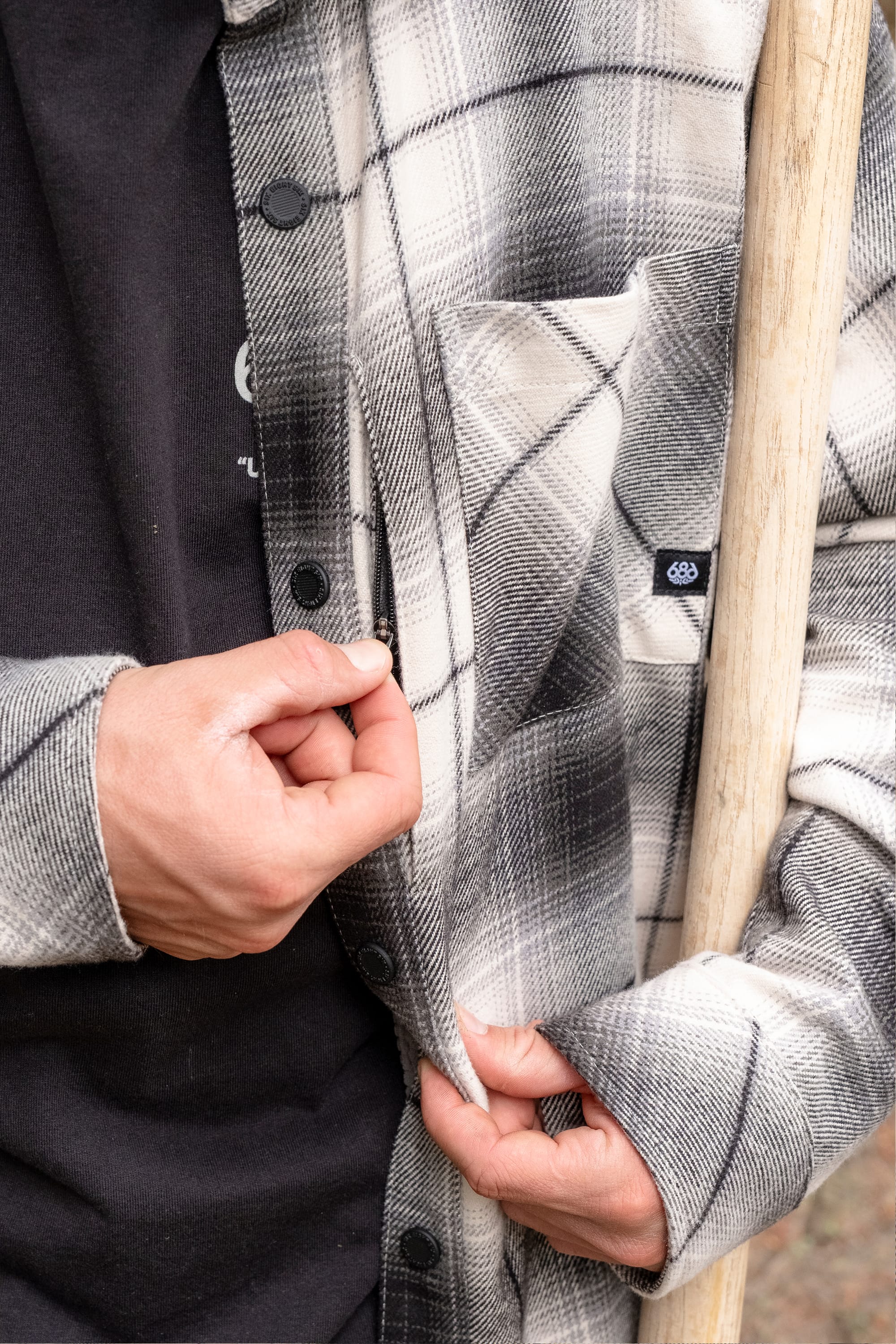 A person wearing the 686 Men's Heat Grid™ Flannel by 686 holds the edge of their shirt with one hand and a wooden stick with the other.