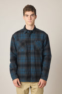 A young man stands facing forward in the 686 Men's Heat Grid Flannel, a blue and black plaid polyester shirt with two chest pockets by 686, paired with beige pants, set against a plain light background.