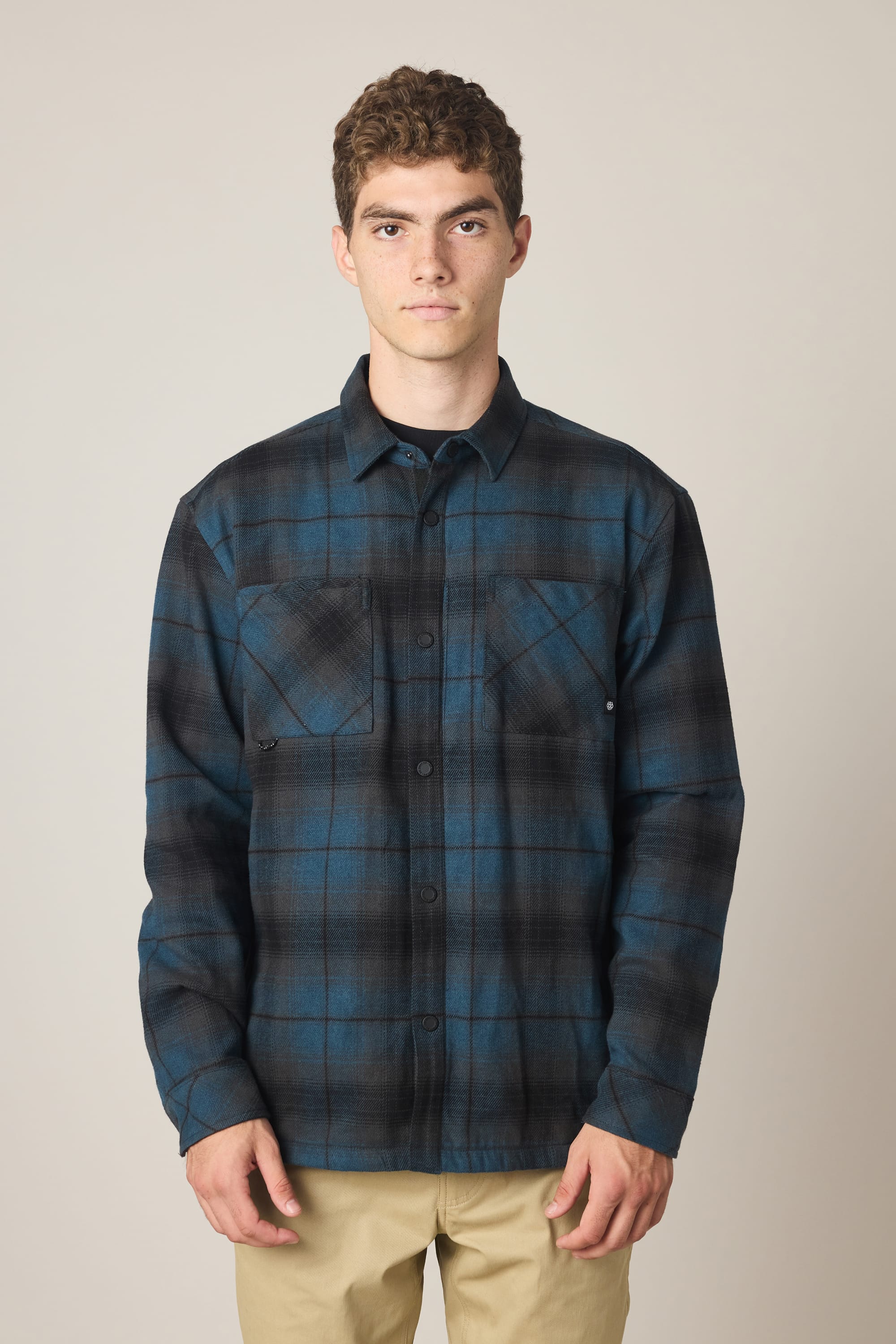A young man stands facing forward in the 686 Men's Heat Grid Flannel, a blue and black plaid polyester shirt with two chest pockets by 686, paired with beige pants, set against a plain light background.