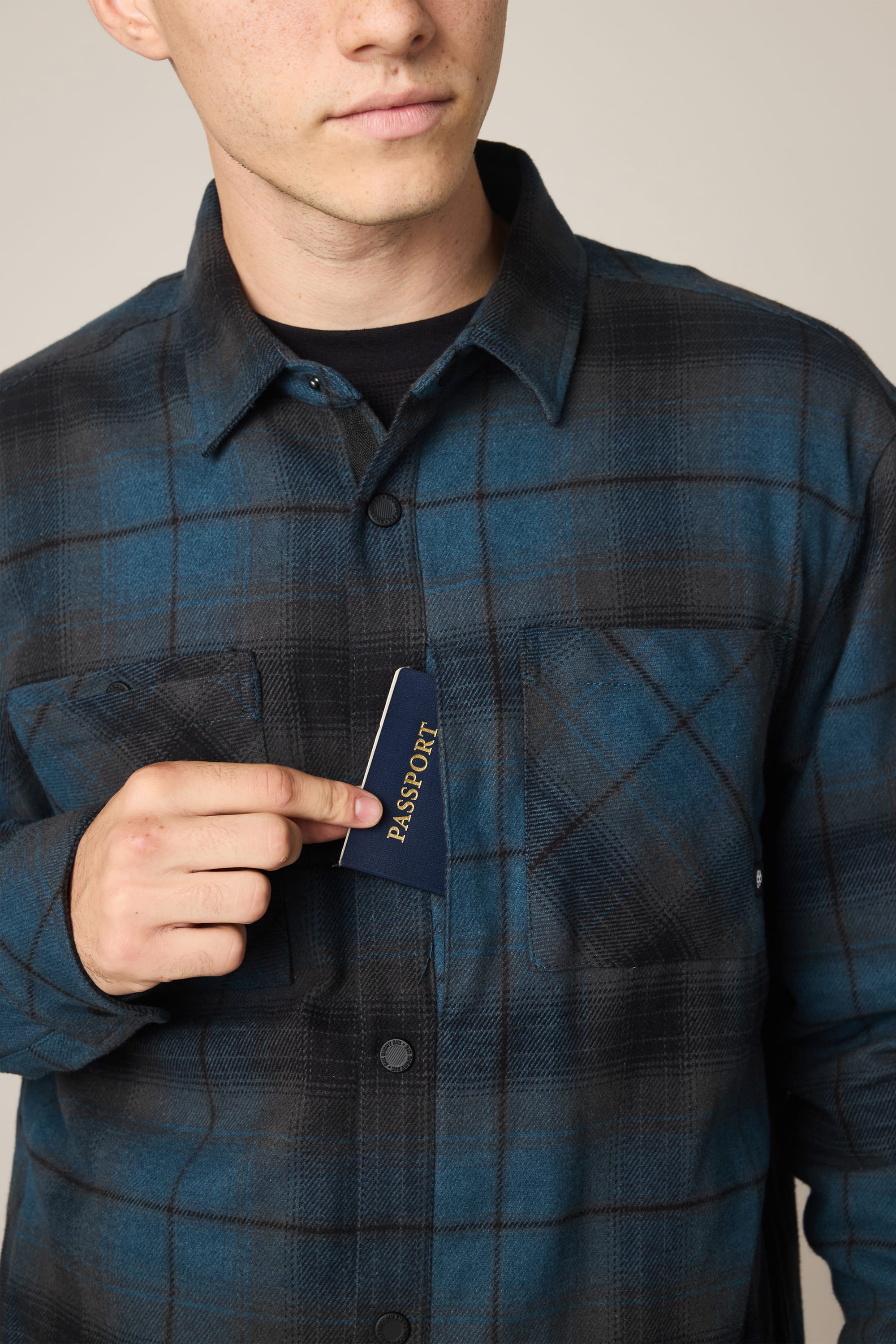 A person in a 686 Men's Heat Grid Flannel places a navy passport with gold lettering into their chest pocket, showing only the lower part of their face.