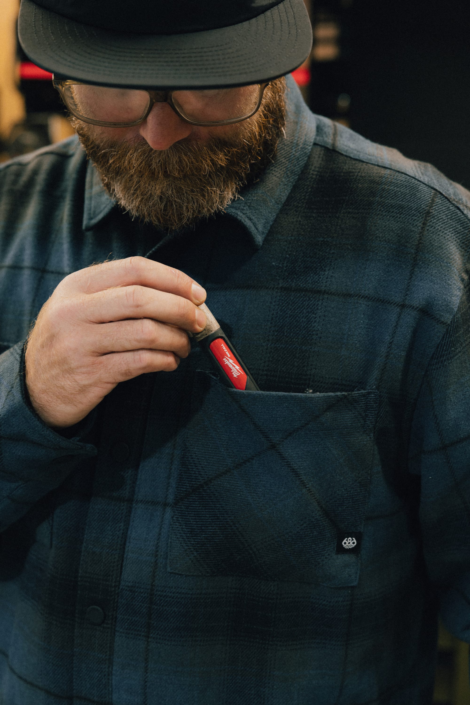 A bearded man wearing glasses and a 686 Men's Heat Grid™ Flannel shirt places a red Swiss Army knife into his chest pocket.