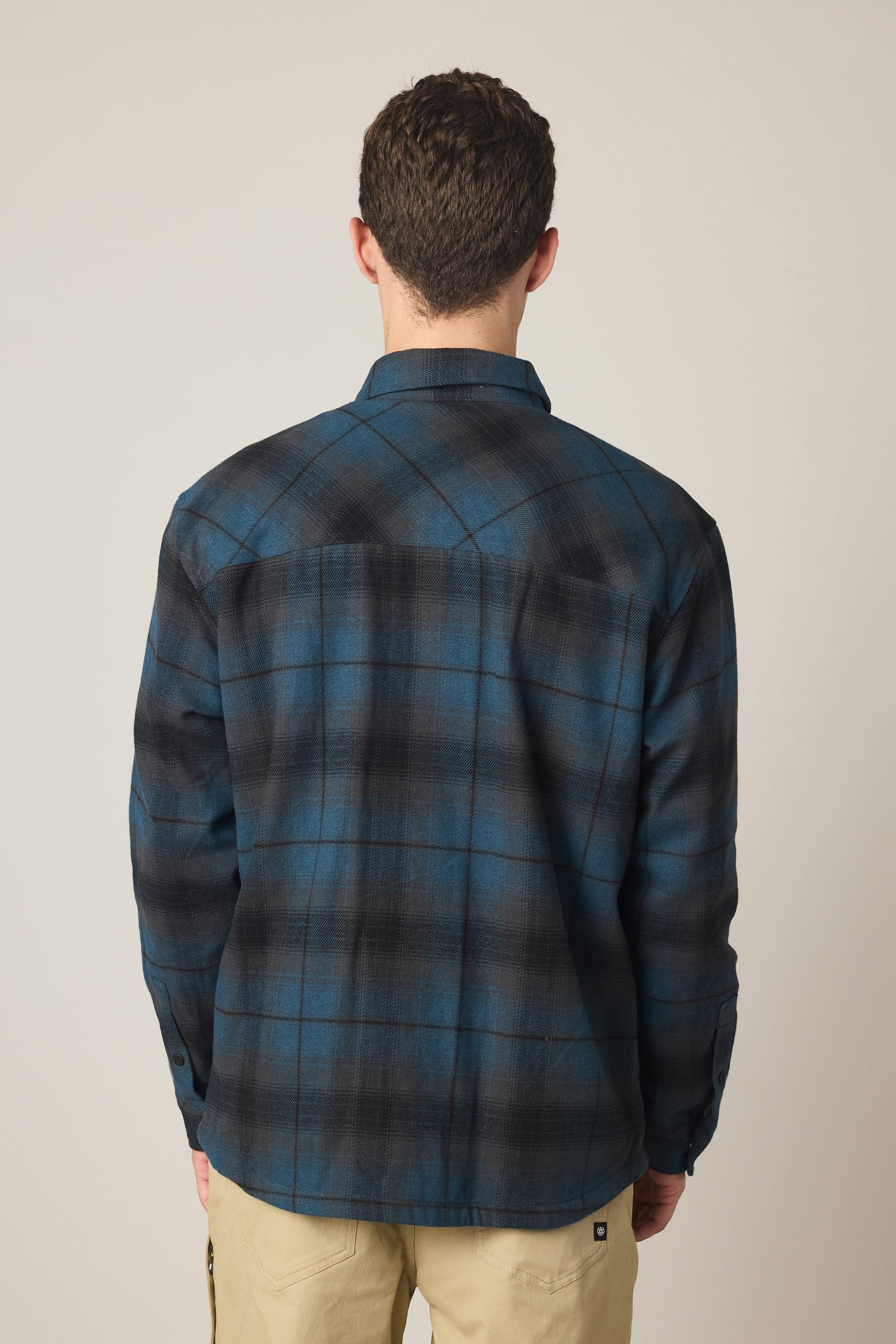 A man stands facing away, wearing the 686 Men's Heat Grid™ Flannel in blue and black plaid and light khaki pants against a plain light background.