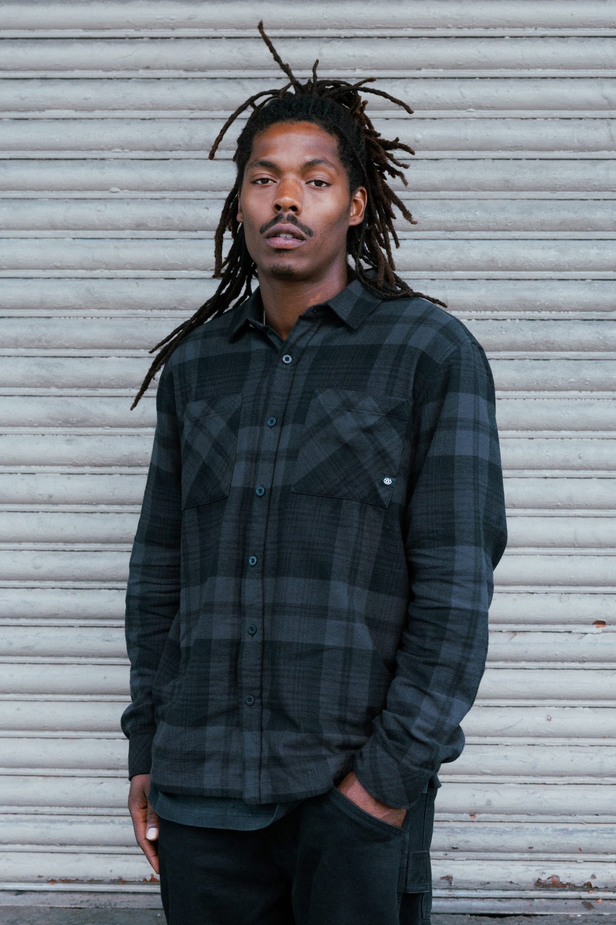 A man with dreadlocks wearing a dark grey and black plaid 686 Men’s SMARTY® Headwall Flannel midweight cotton shirt.
