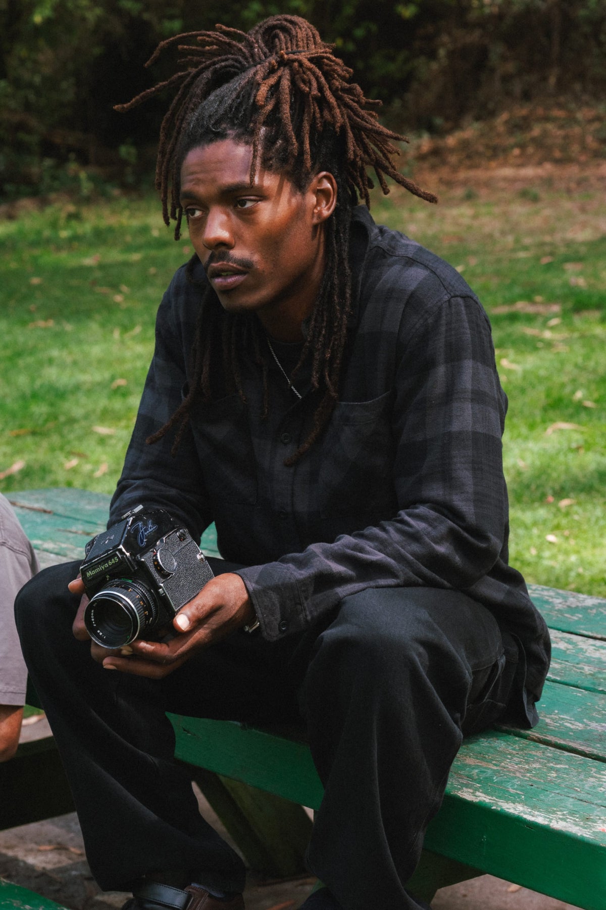 A person wearing a dark grey plaid 686 Men’s SMARTY® Headwall Flannel sits on a park bench holding a vintage medium format camera.