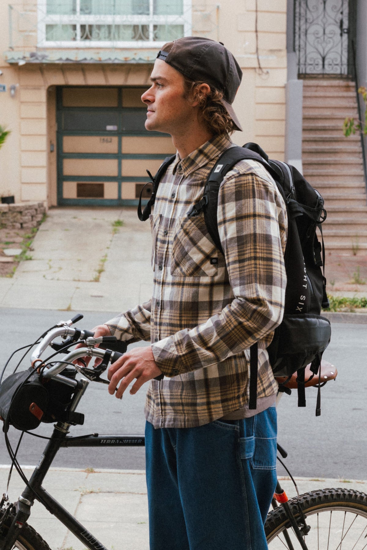 A person wearing a tan and grey plaid 686 Men’s SMARTY® Headwall Flannel, a baseball cap, and a black backpack, standing next to a bicycle on a residential street.
