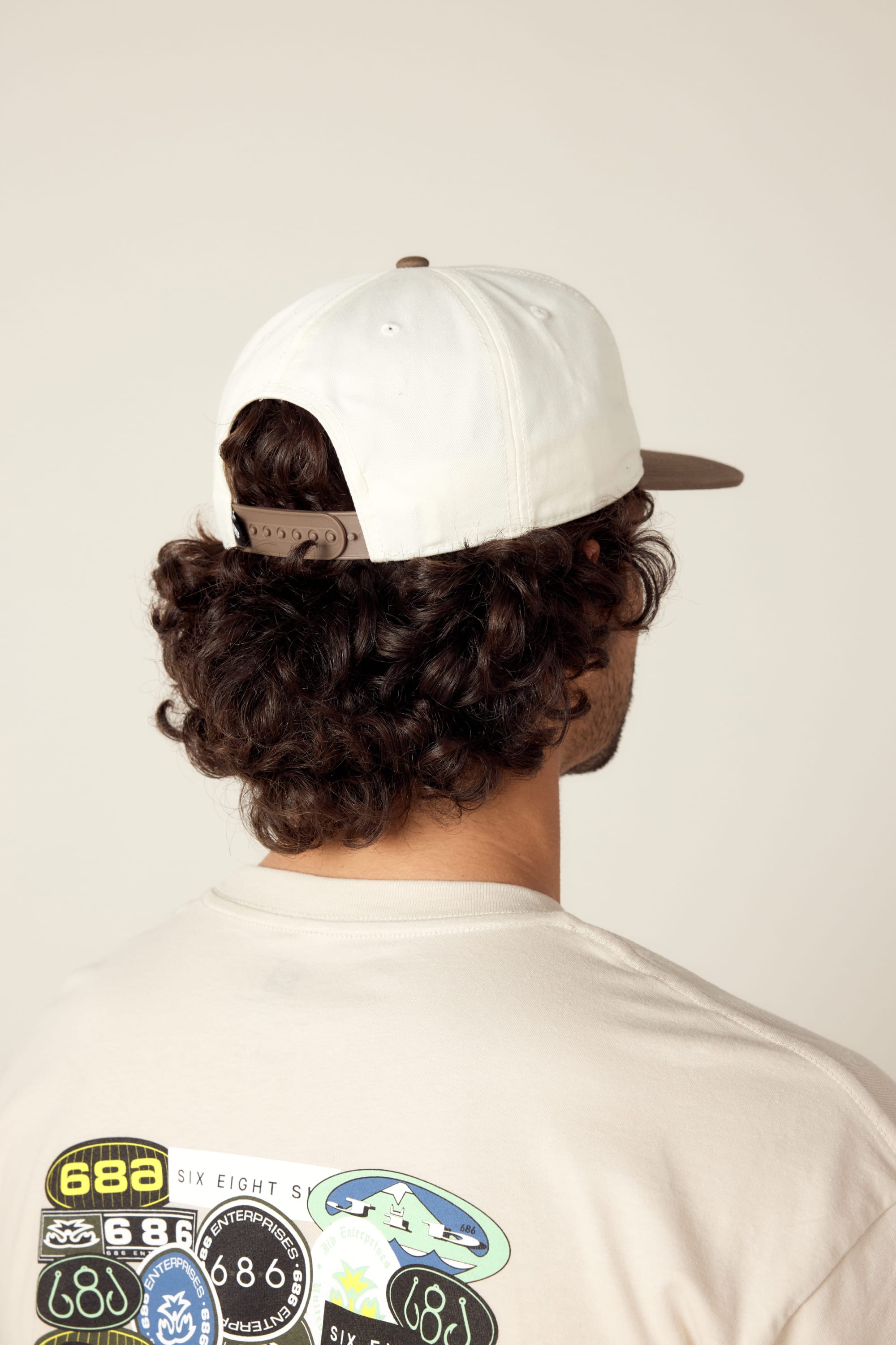 A person with curly hair wears the 686 Cross-Cut Unstructured Hat backwards and a beige T-shirt with colorful graphic patches on the back, shown from behind against a neutral background.
