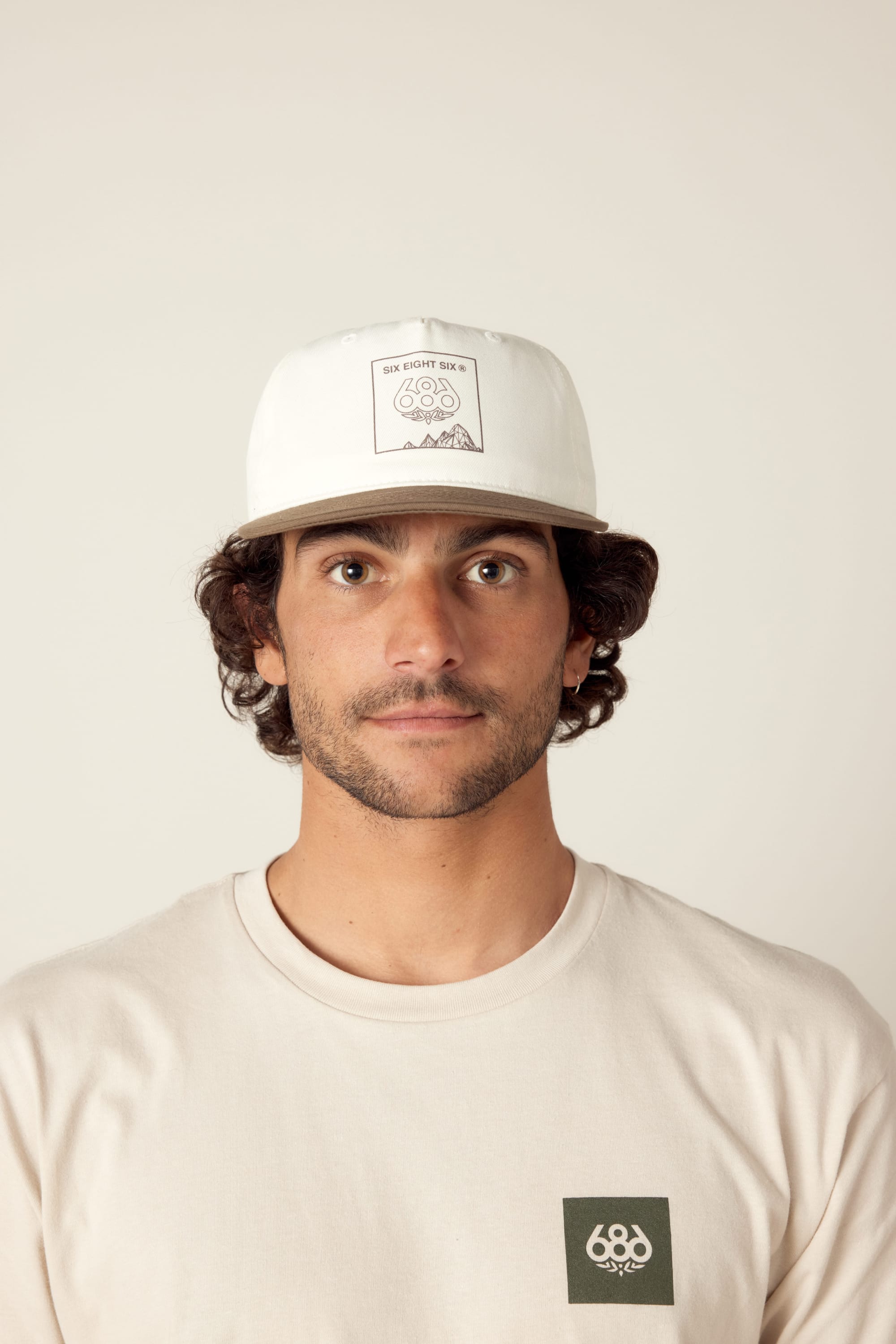 A man with curly brown hair wears the 686 Cross-Cut Unstructured Hat and a cream-colored t-shirt with a 686 logo patch, looking directly at the camera against a plain light background.