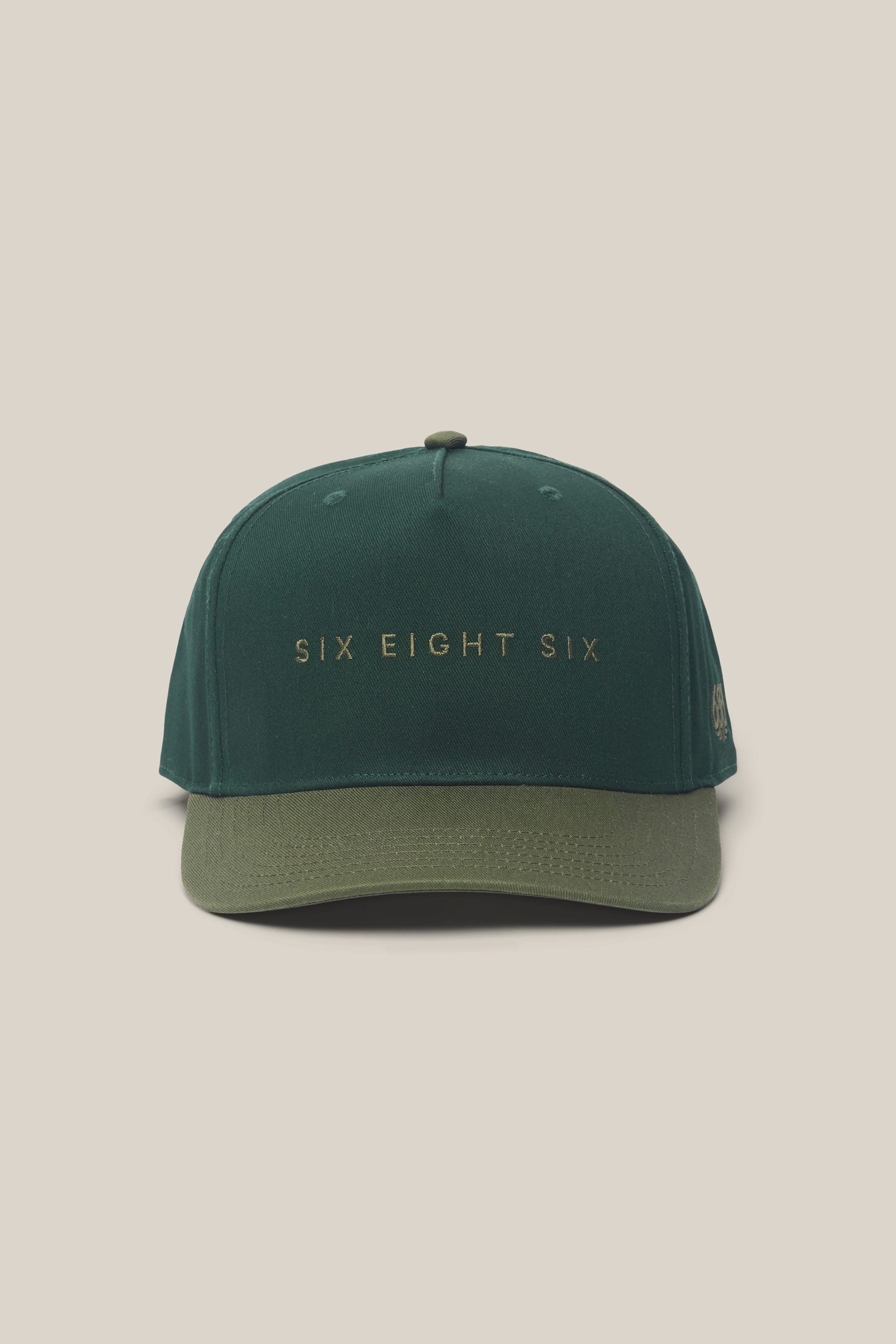 The 686 High-Pro Ses Two Tone Structured 5 Panel Hat by 686 is a men's headwear piece crafted from dark green woven cotton with a slightly lighter green brim. The front displays "SIX EIGHT SIX" embroidered in gold letters, and the background is set against a plain off-white color.