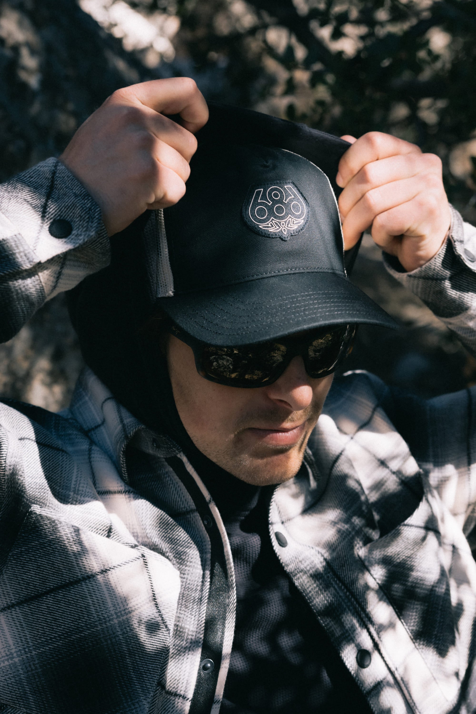 A person wearing sunglasses, a plaid jacket, and the 686 Mid-Pro Wreath Unstructured 5 Panel Mesh Hat by 686 with its circular logo adjusts their hood outdoors in dappled sunlight.