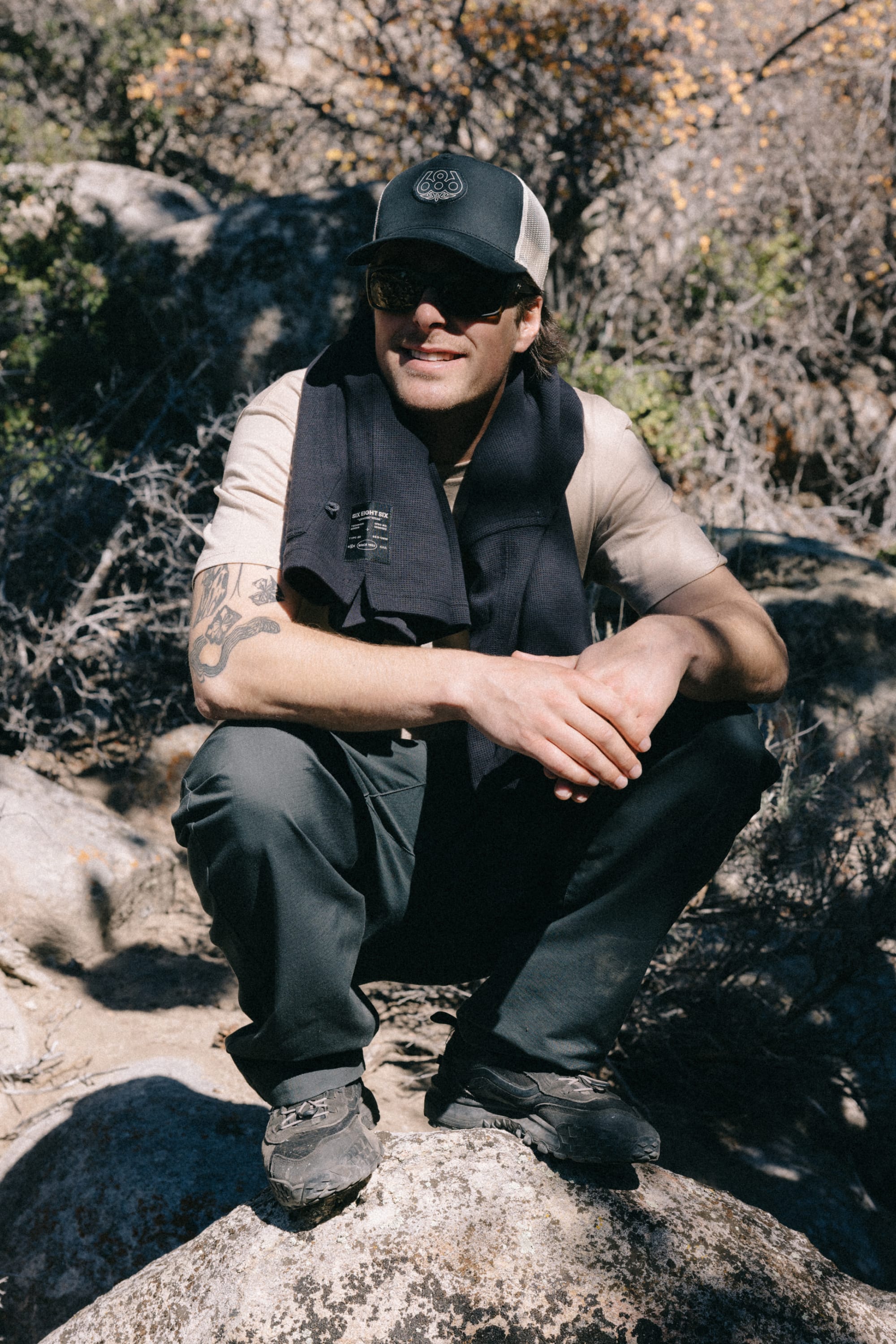 A man wearing sunglasses, outdoor clothing, and a 686 Mid-Pro Wreath Unstructured 5 Panel Mesh Hat by 686 squats on a rock in a sunlit, rocky, and brushy outdoor area.