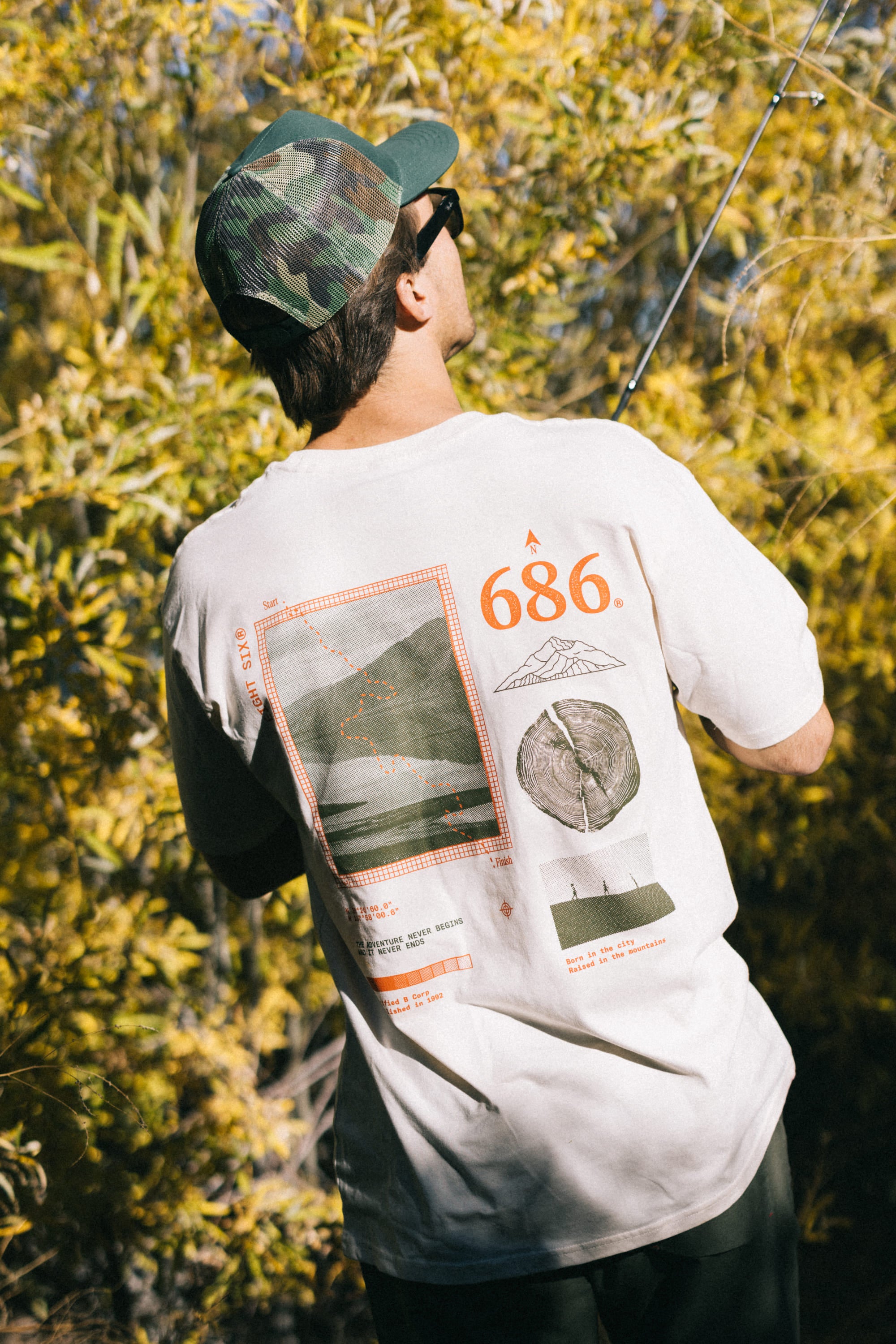 Wearing a white graphic T-shirt and the 686 Mid-Pro Wreath Unstructured 5 Panel Mesh Hat by 686, a person stands outdoors with their back to the camera, holding a fishing rod near leafy yellow-green bushes.